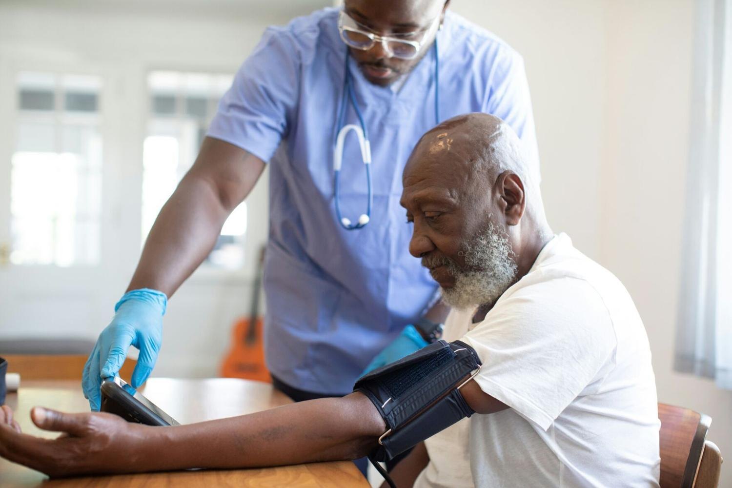 Healthcare worker checking blood pressure of a seated older man using a cuff and tablet.