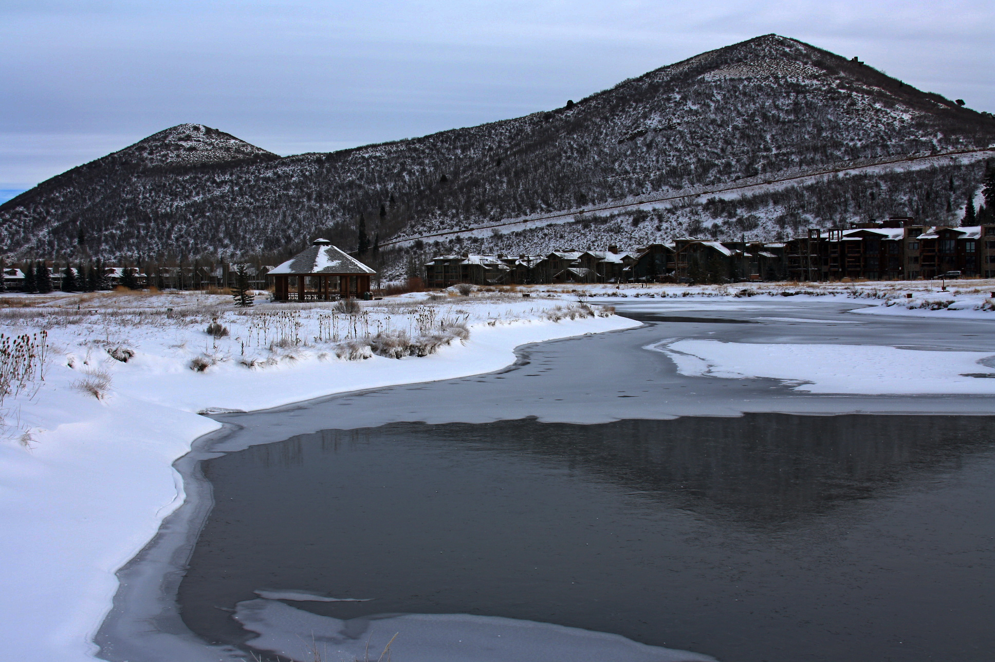 Partially frozen pond with a gazebo and snow-dusted mountain peaks at a ski resort.