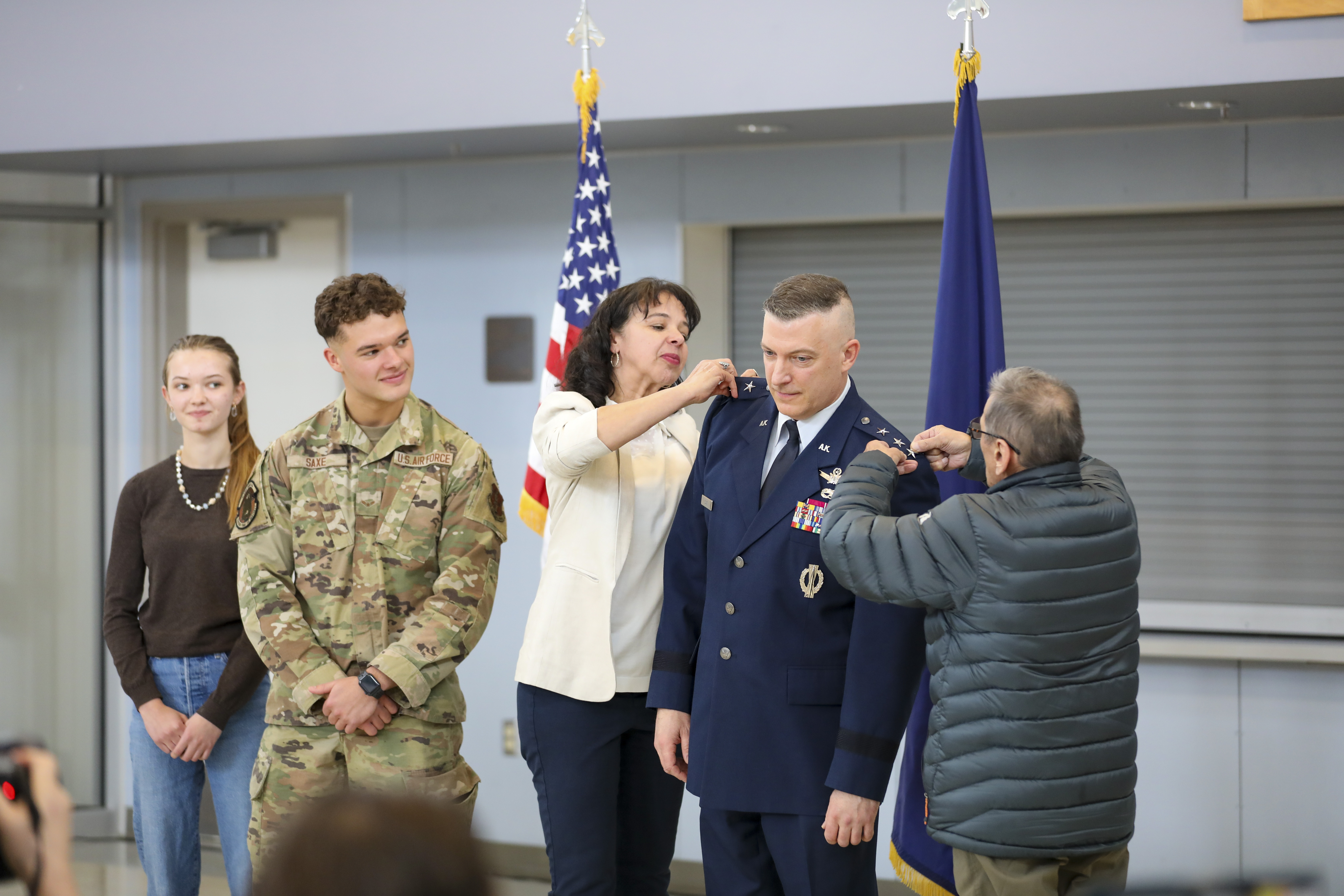 Two people simultaneously pin rank insignia onto a uniformed officer's shoulders at an indoor military ceremony.