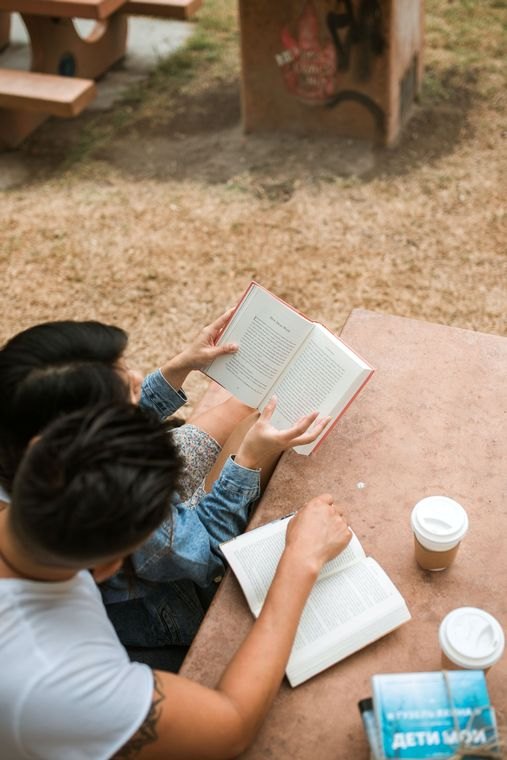 Two people reading open books at an outdoor table, viewed from above.