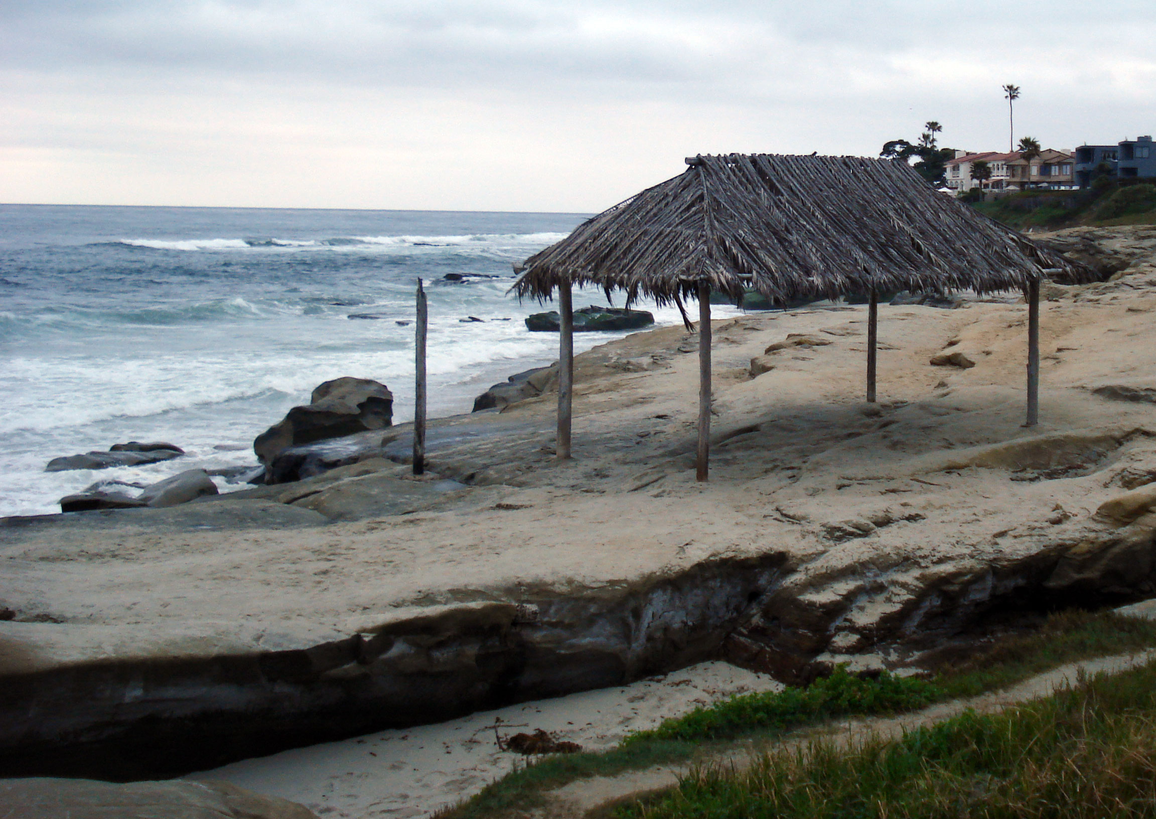 Thatched-roof palapa structure on a sandy beach with rocky shoreline and breaking ocean waves.