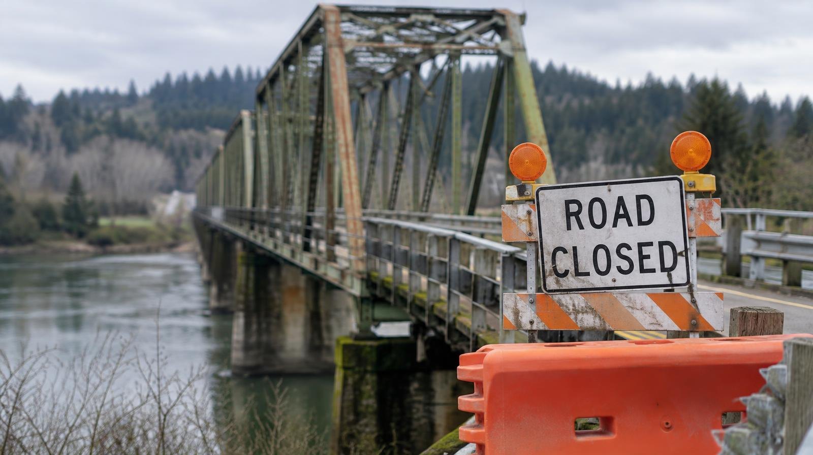 A weathered steel truss bridge over a river with orange safety barriers blocking the entrance.