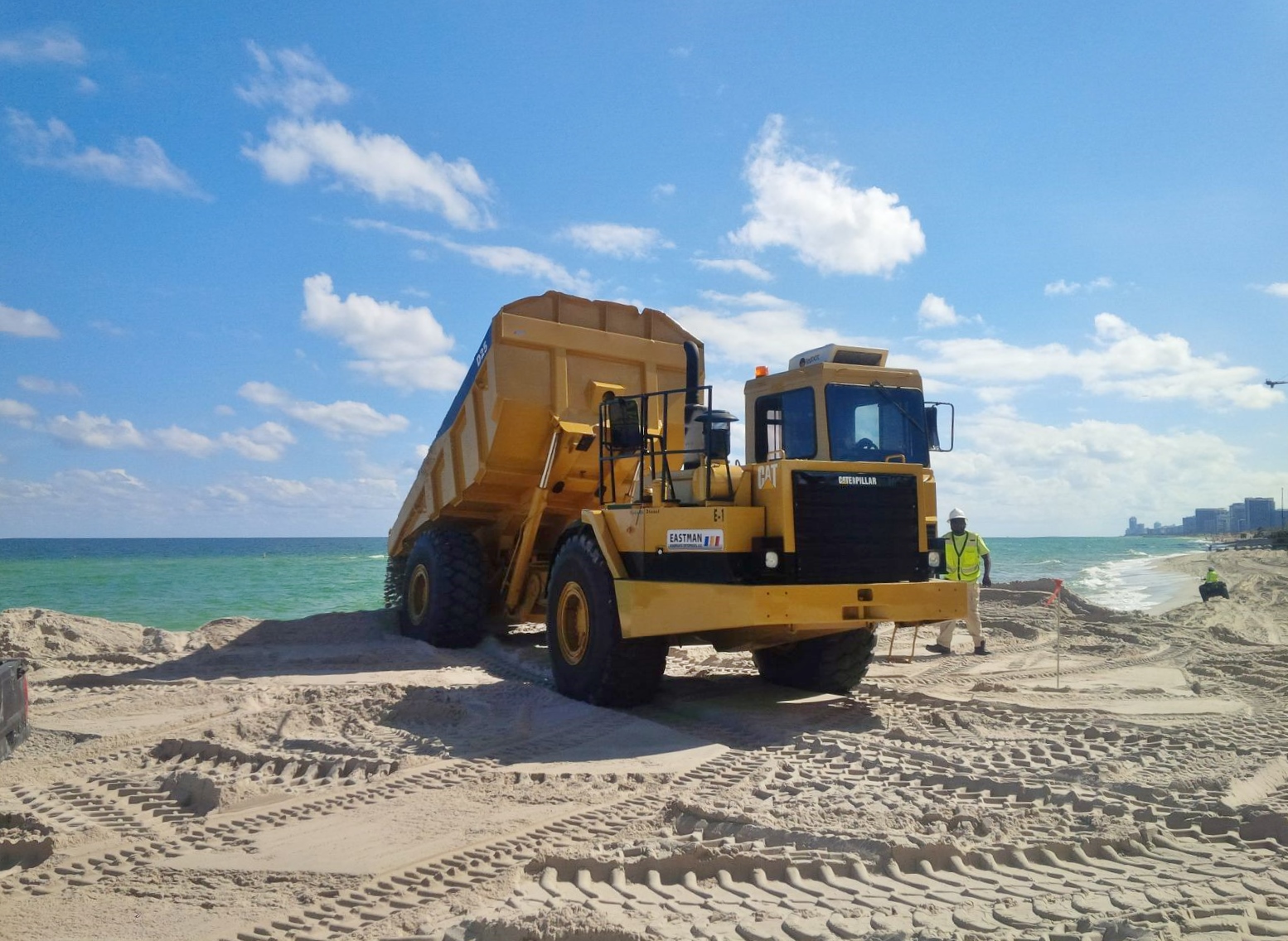 Yellow dump truck depositing sand on a beach near a pier during renourishment work.