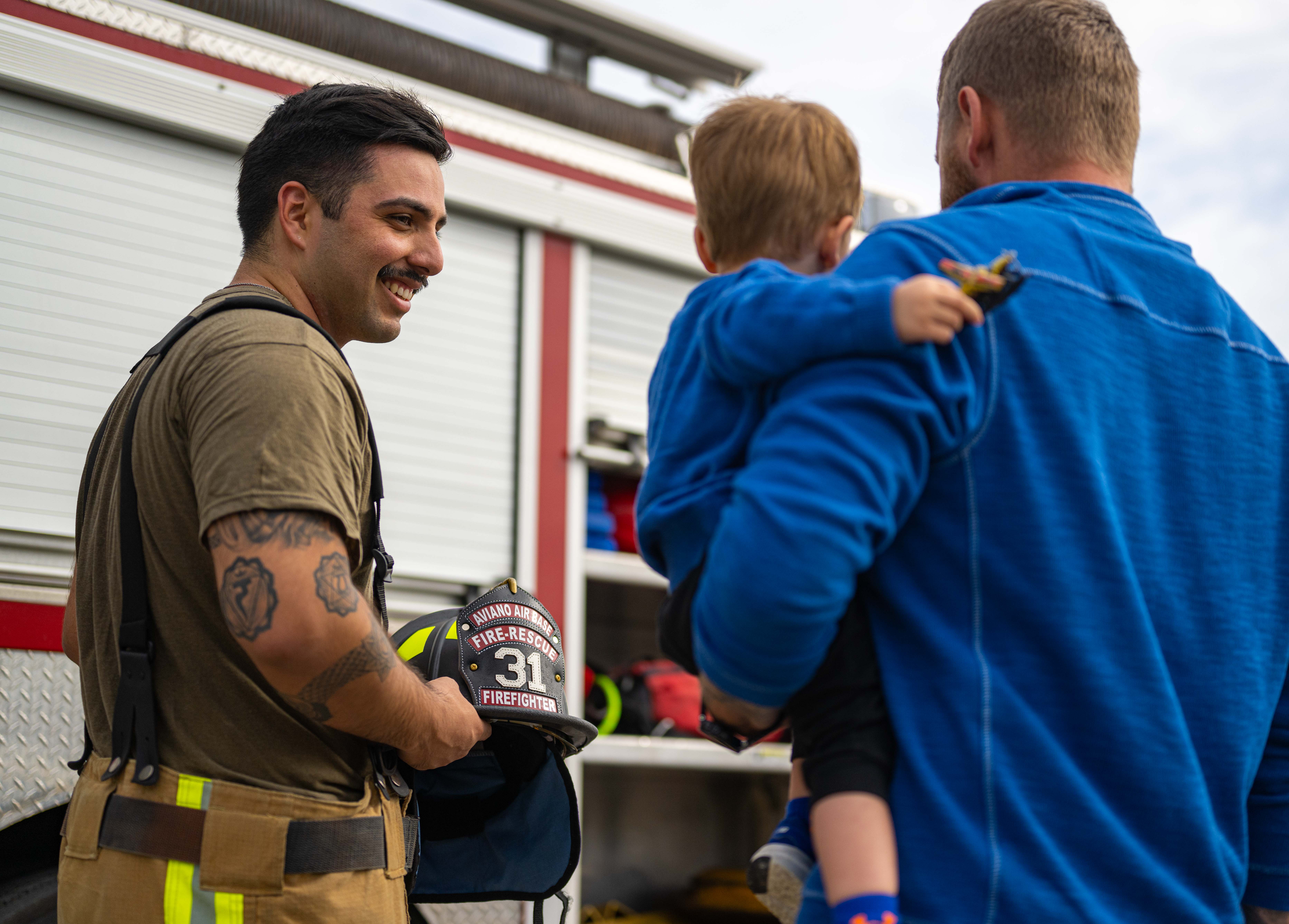 A firefighter in turnout gear smiles and holds a helmet near a fire truck while talking with a man holding a child.