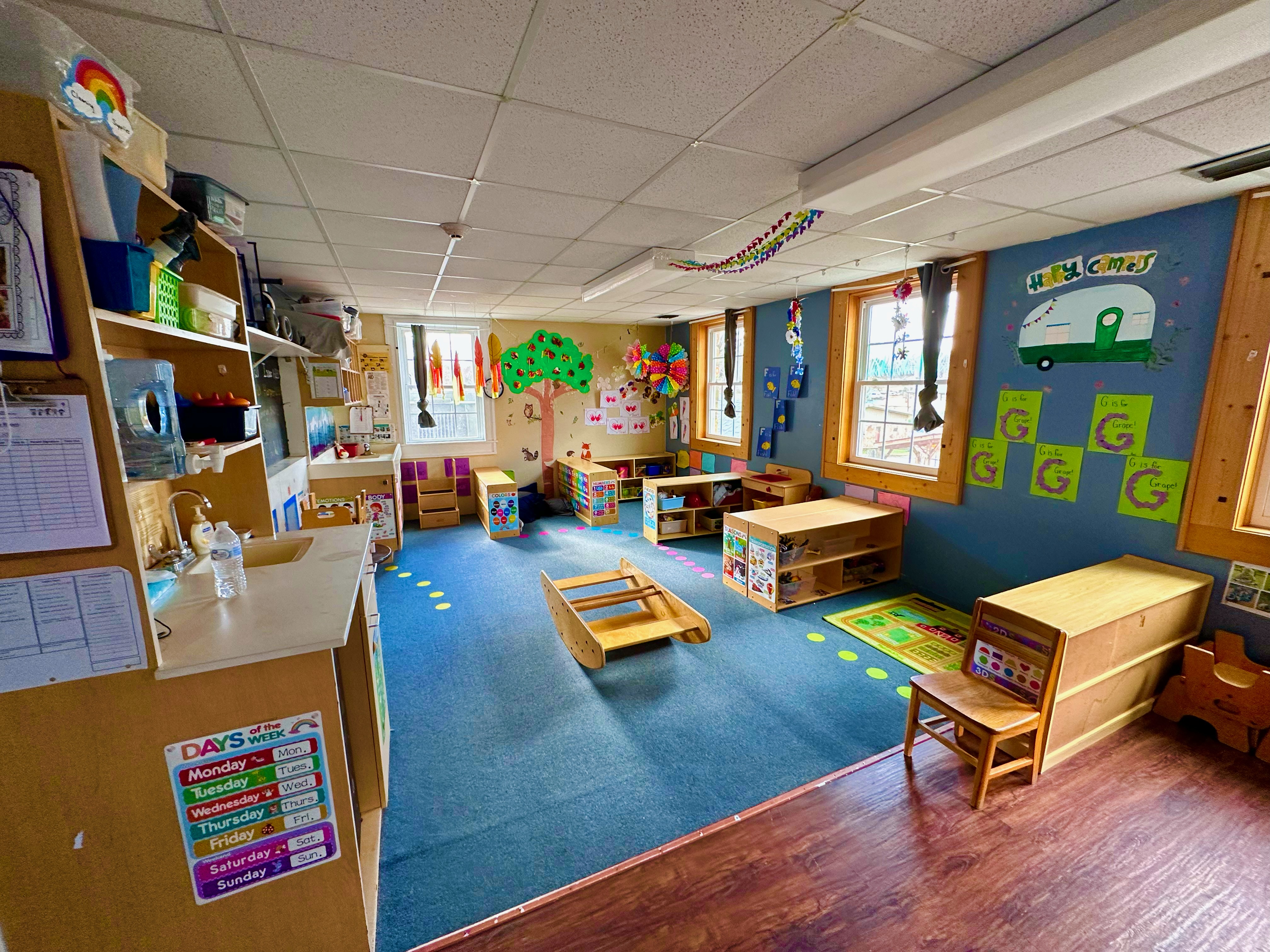 Brightly decorated daycare classroom with child-sized furniture, colorful wall art, and a blue carpet.