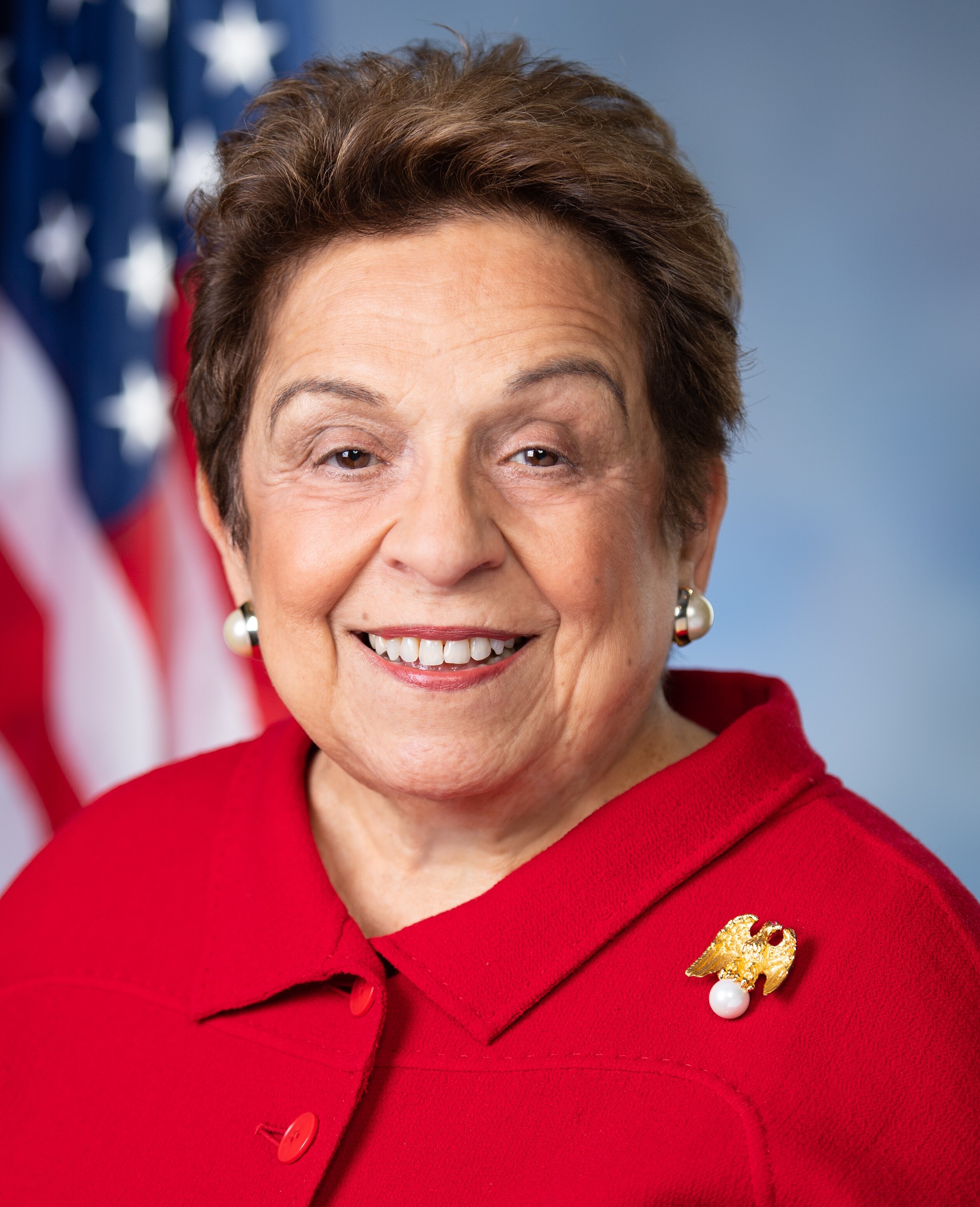 Official portrait of Donna Shalala wearing a red blazer with a gold brooch in front of an American flag.