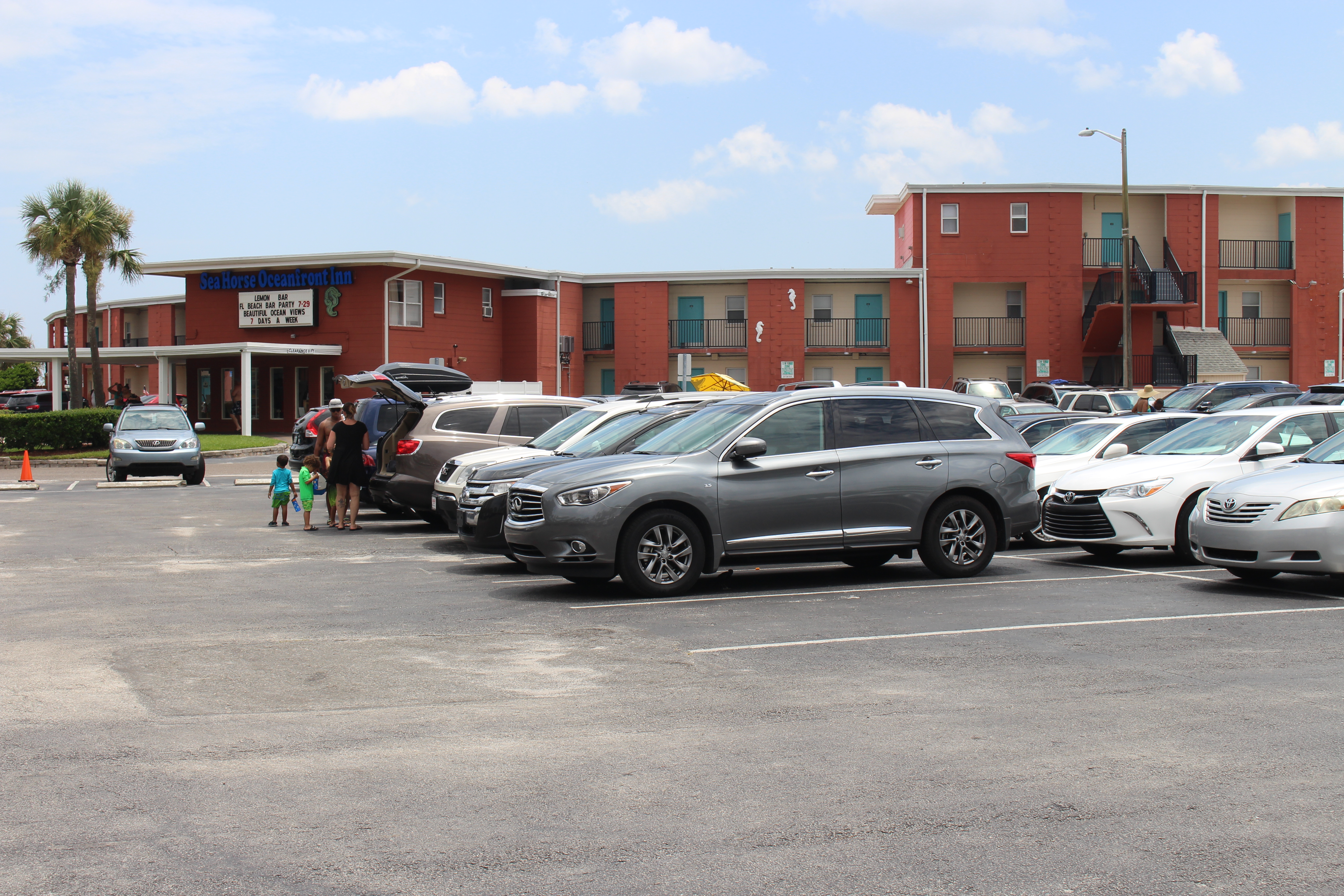 Two-story red-brick inn with palm trees and parked cars on a sunny day.