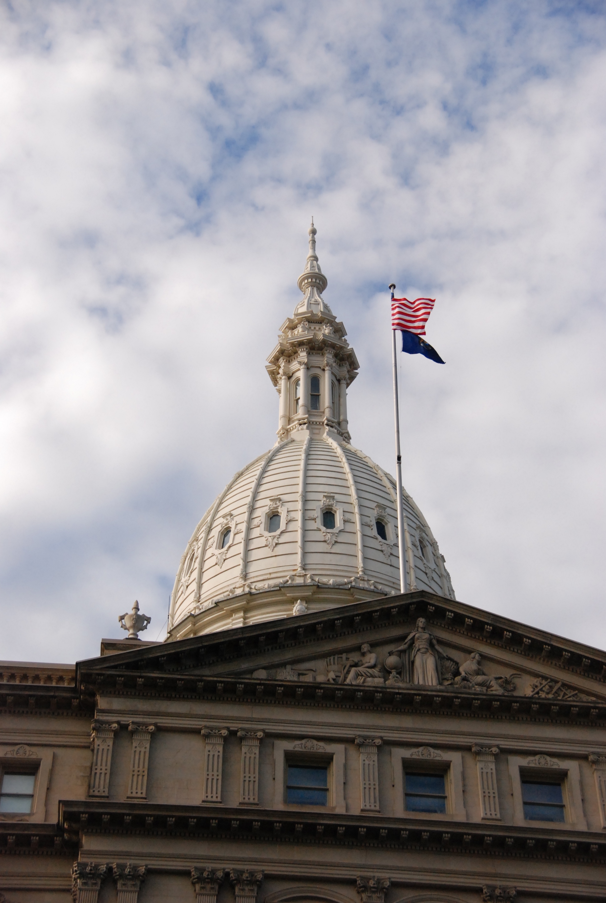 Michigan State Capitol dome and flags viewed from below against a partly cloudy sky.