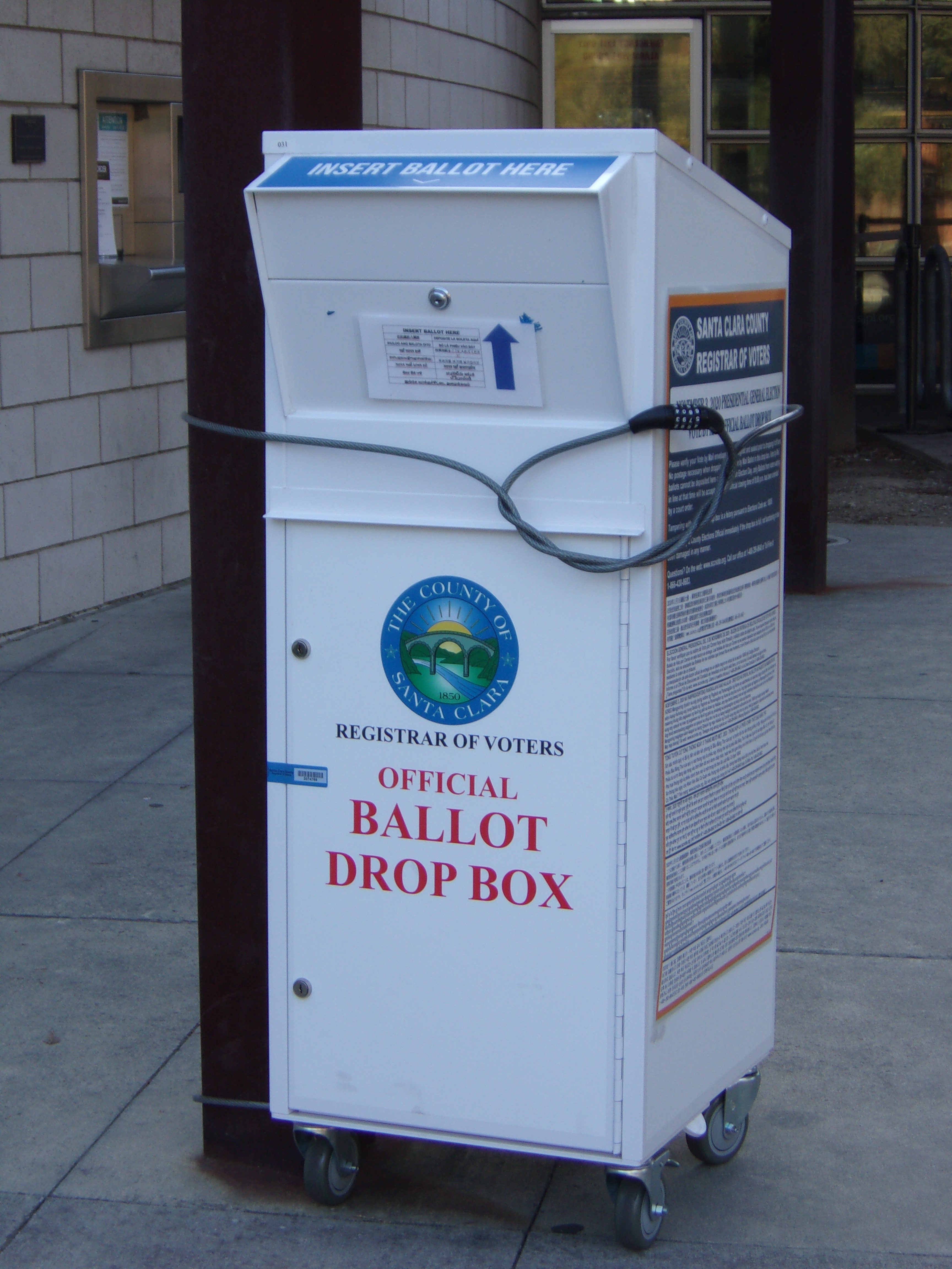 White official ballot drop box on wheels labeled Santa Clara County Registrar of Voters, outdoors on a sidewalk.