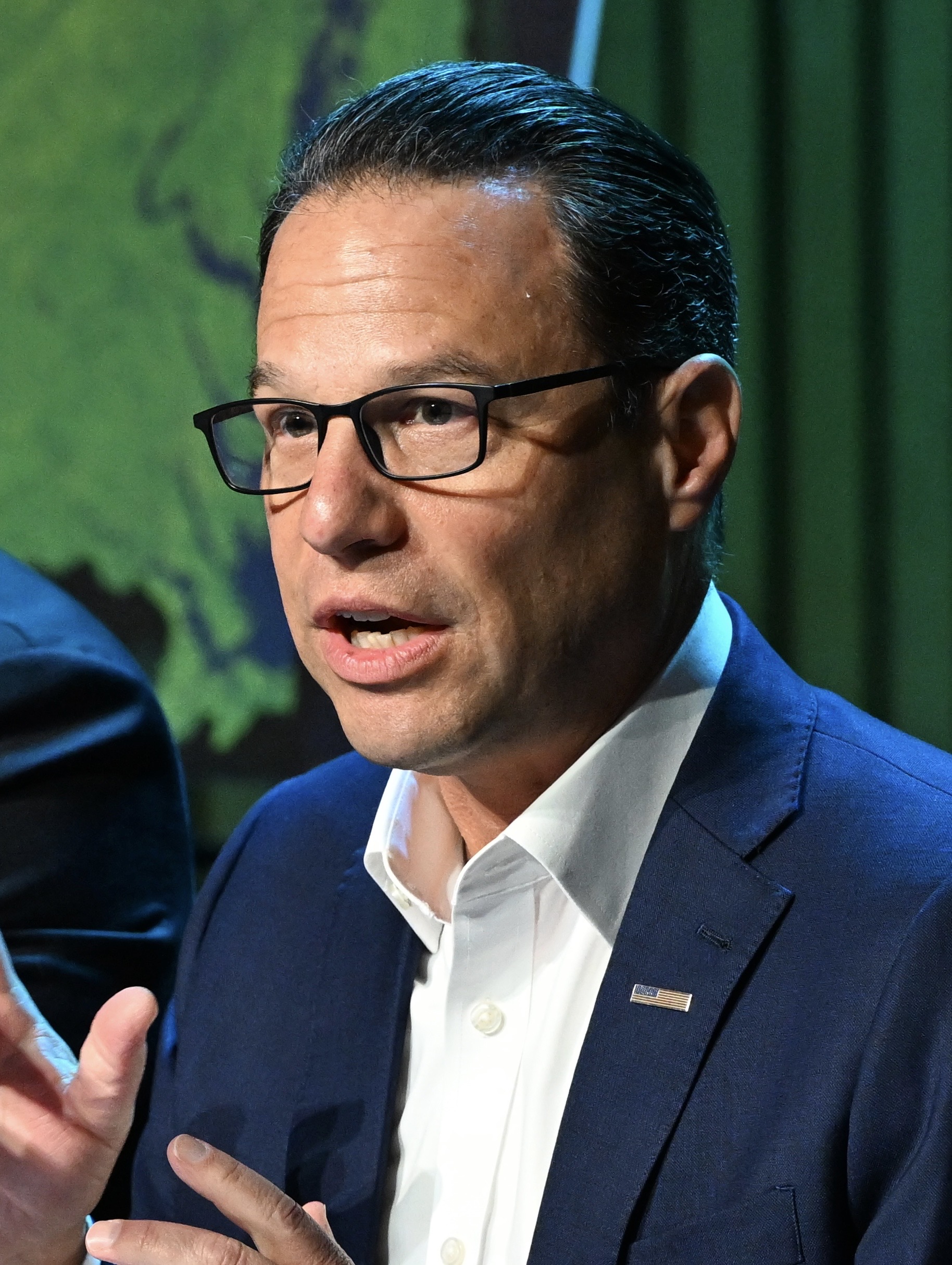 Man in navy blazer and dark-framed glasses gesturing while speaking at an event.