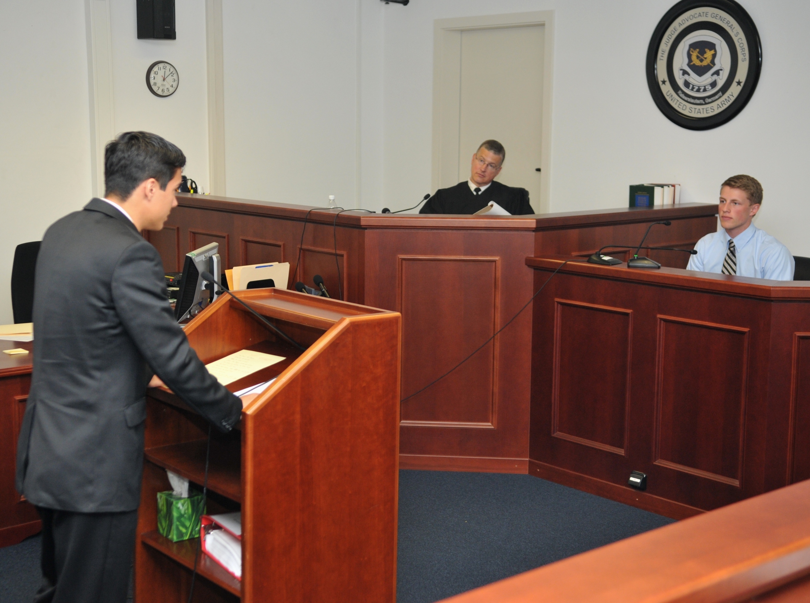 A student prosecutor stands at a courtroom podium as a robed judge presides at the bench.
