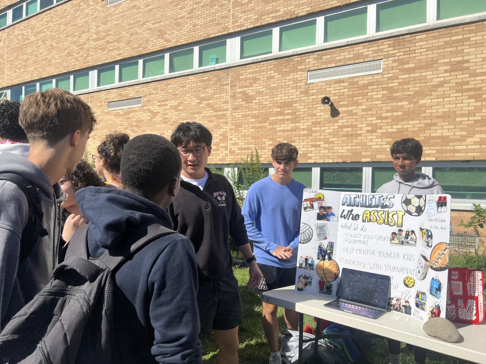 Students gathered around an outdoor table with a trifold display board at a school activities fair.