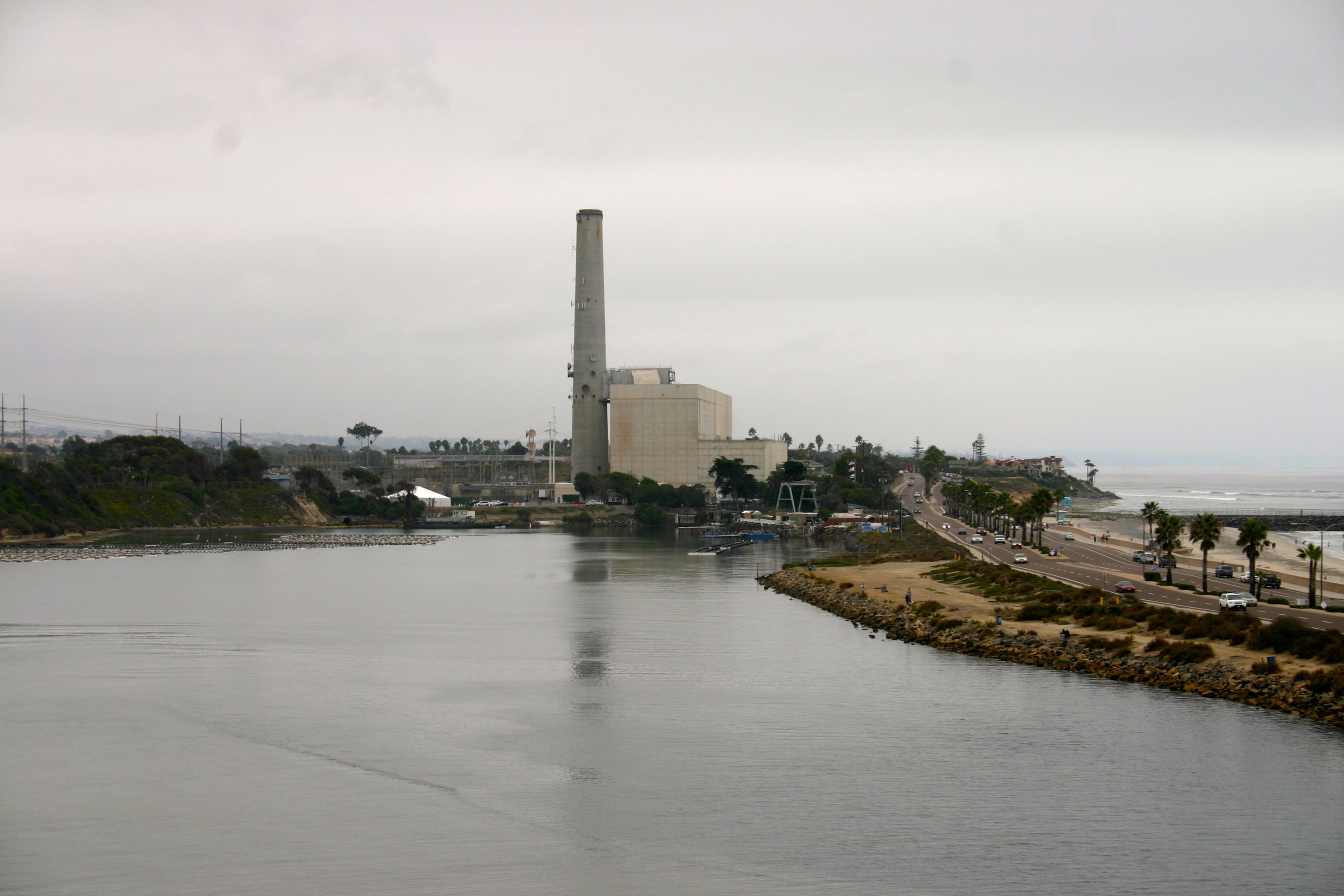 Industrial power station with tall smokestack reflected across a calm coastal lagoon.
