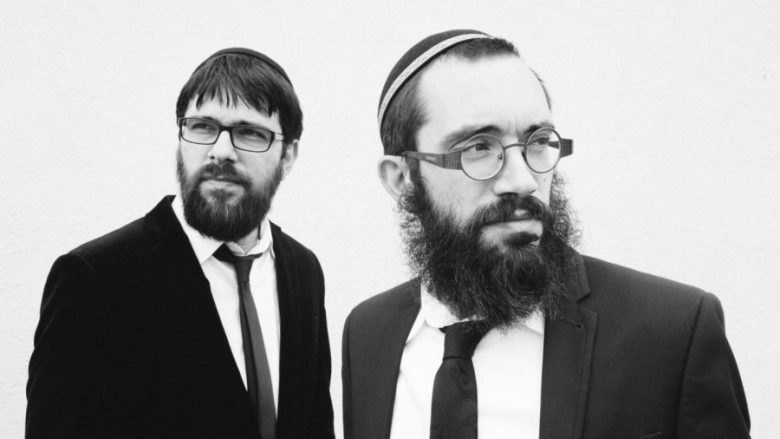 Two bearded men in dark suits and kippot pose in a black-and-white portrait.
