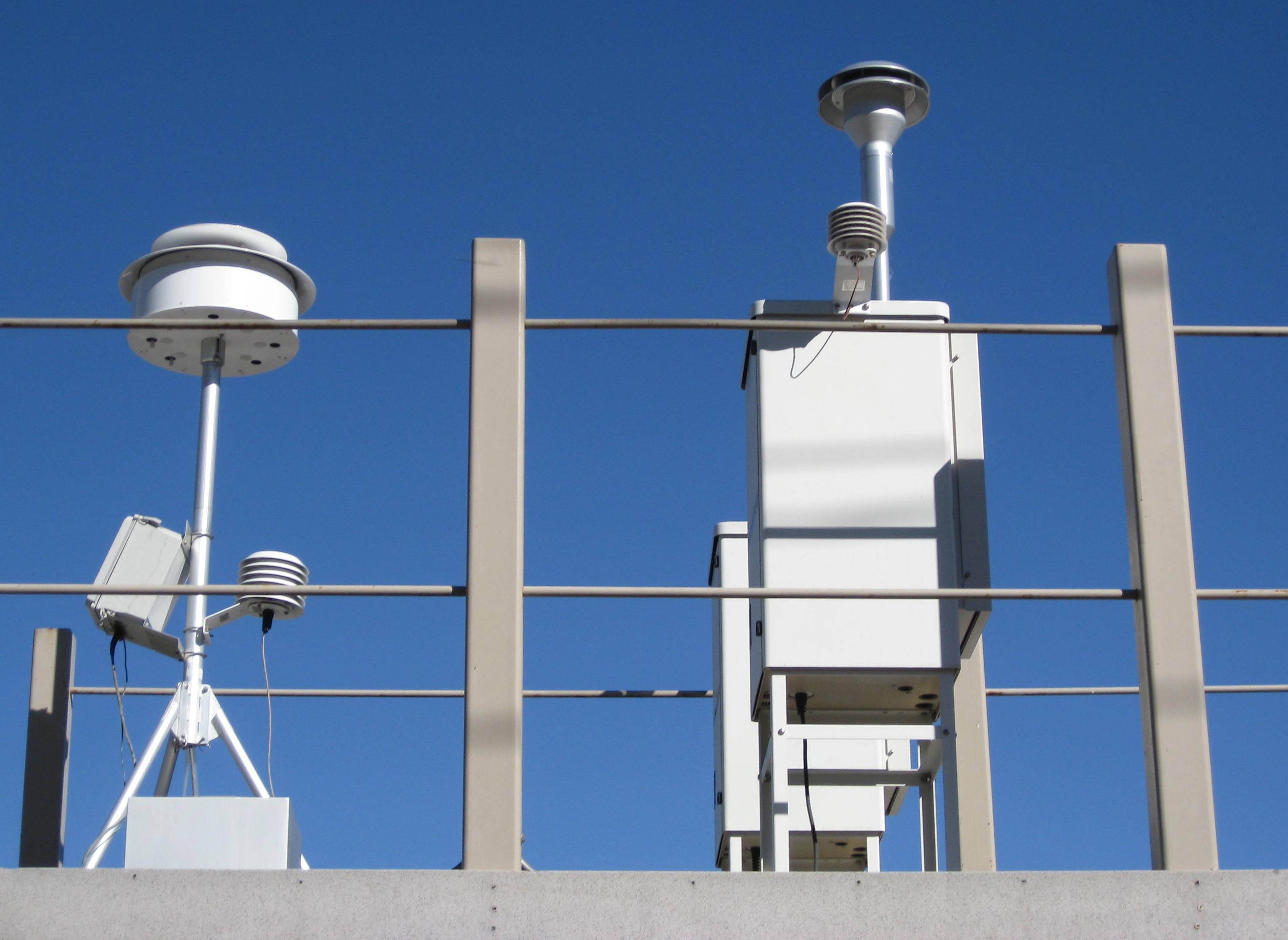 Rooftop air quality monitoring sensors mounted on a metal railing against a blue sky.