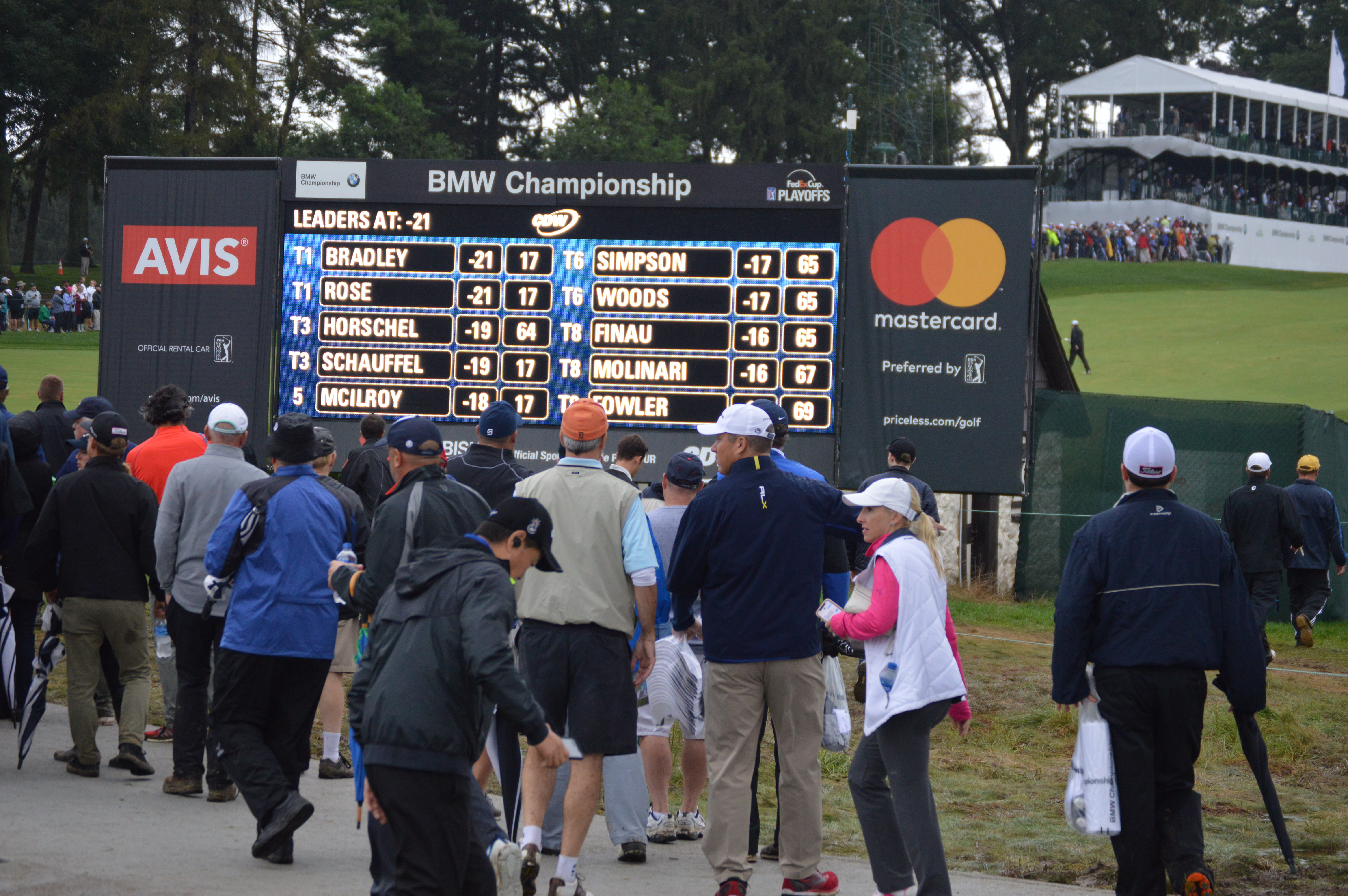 Crowd of spectators standing near a large golf tournament leaderboard.
