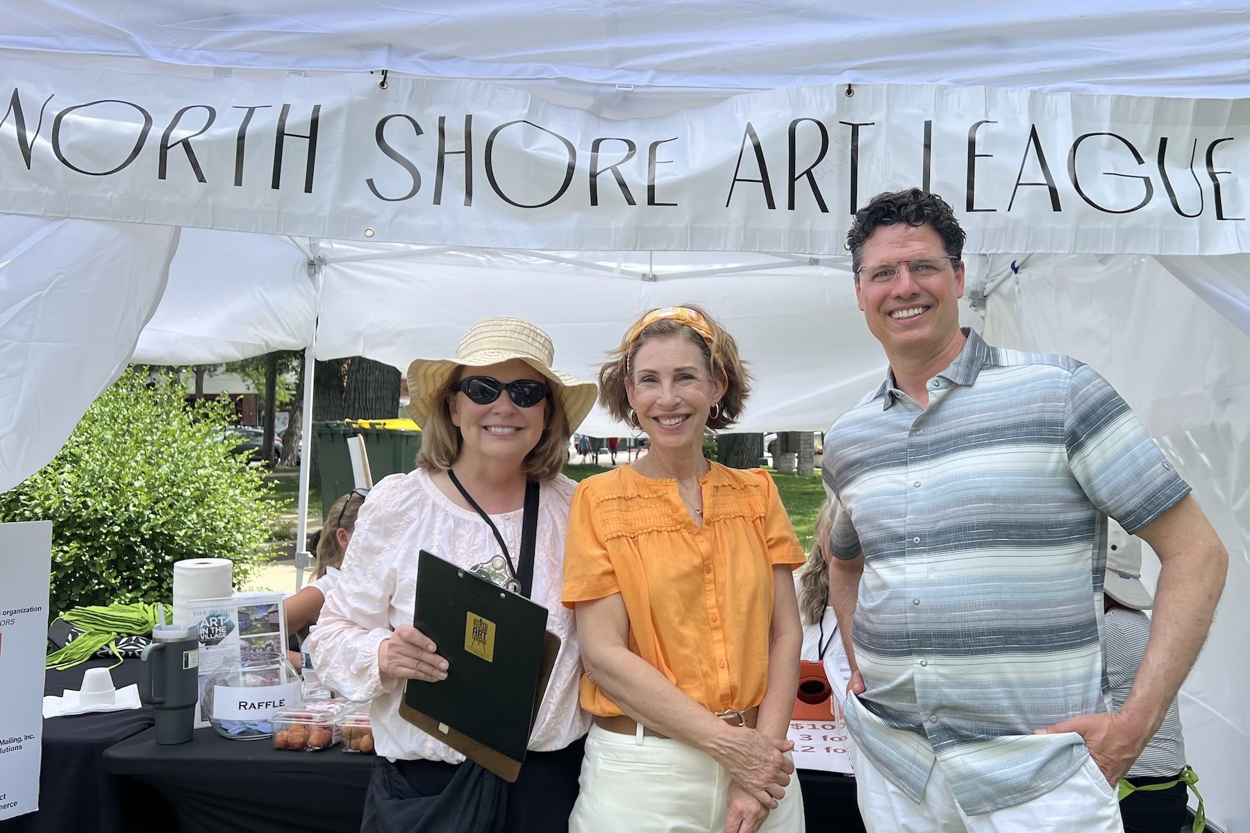 Three people posing under a white tent banner reading North Shore Art League at an outdoor event.