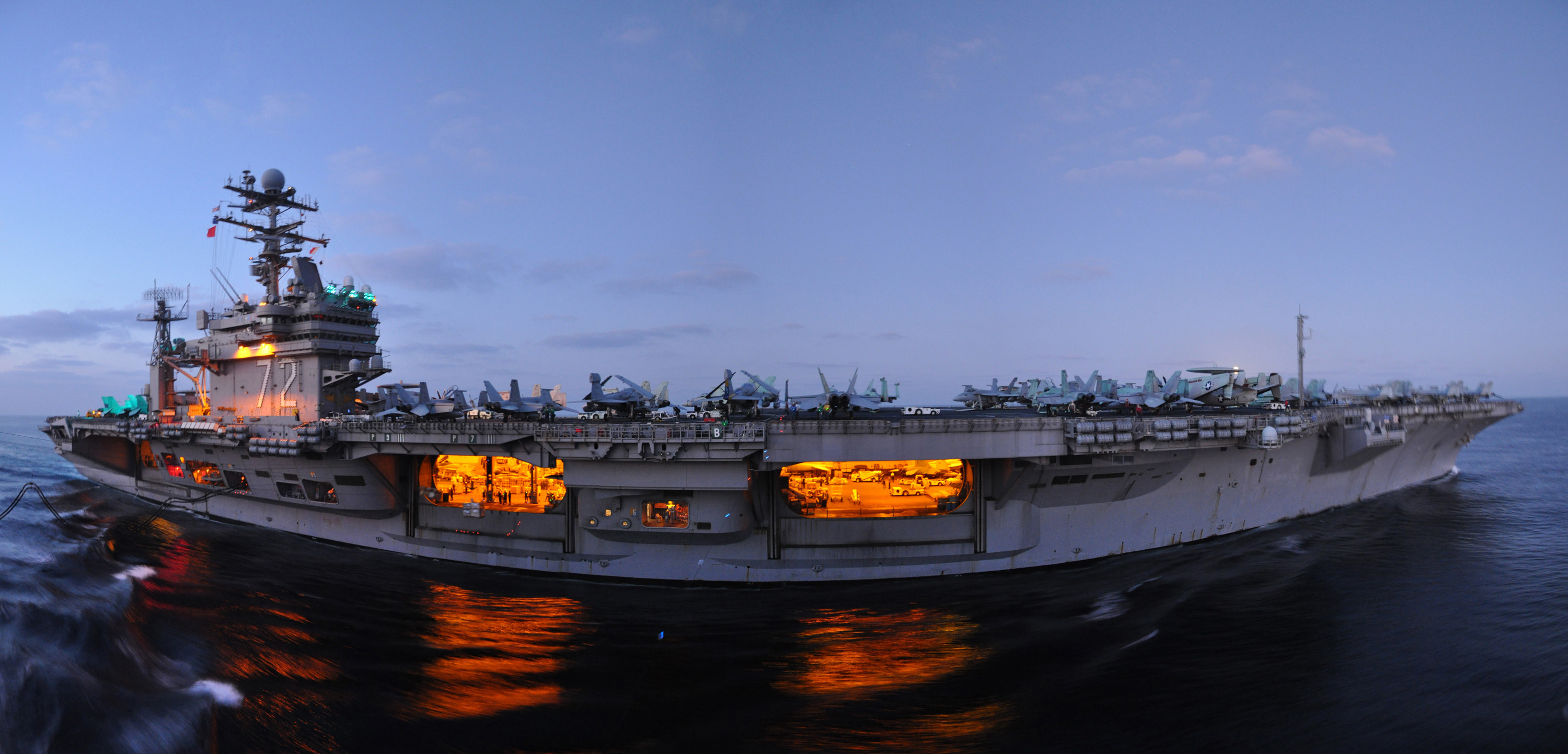 Aircraft carrier at sea at dusk, with an illuminated hangar bay and aircraft visible on the flight deck.