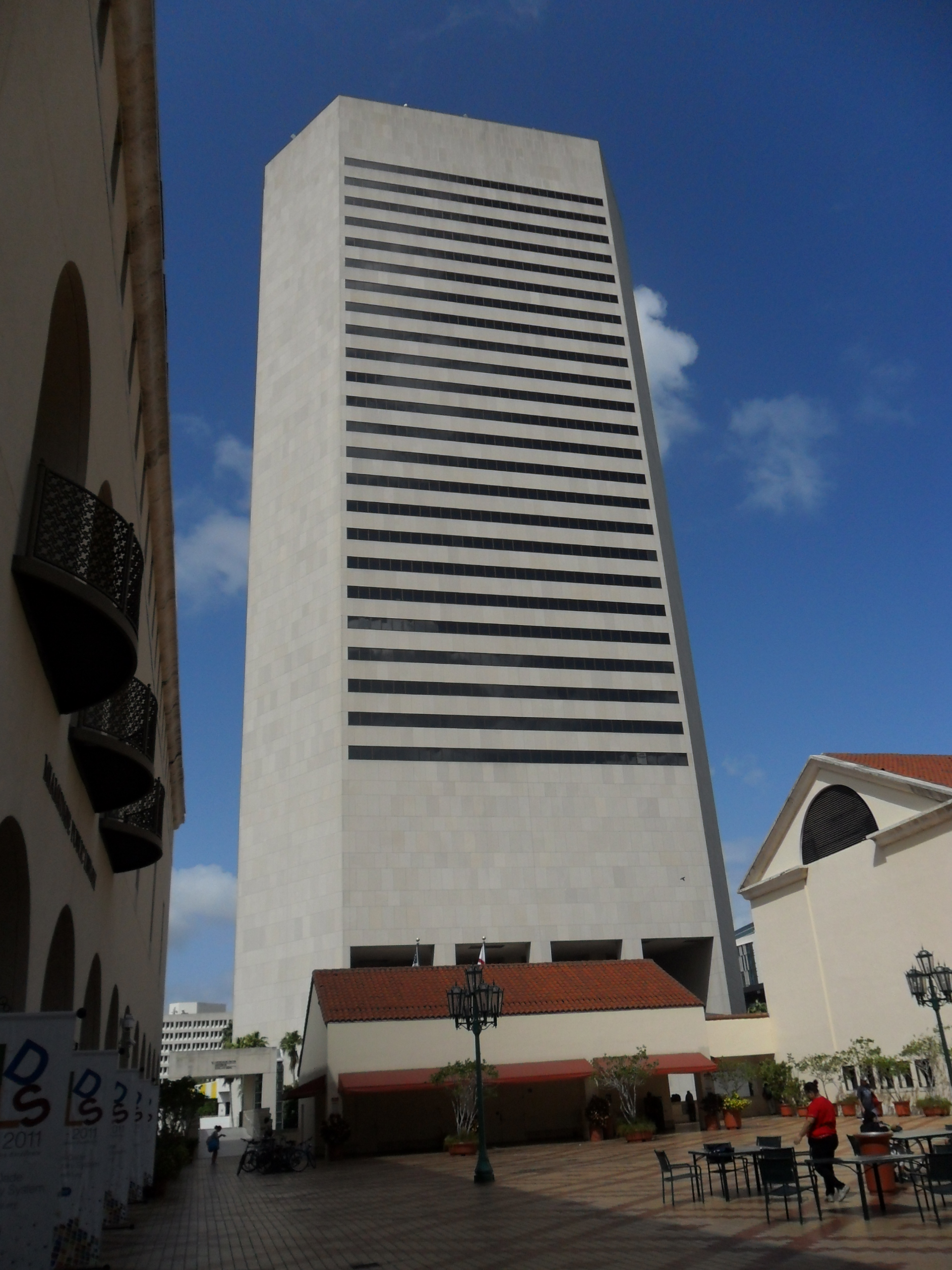 Tall white modernist government tower viewed from a plaza with outdoor seating and a lamp post.