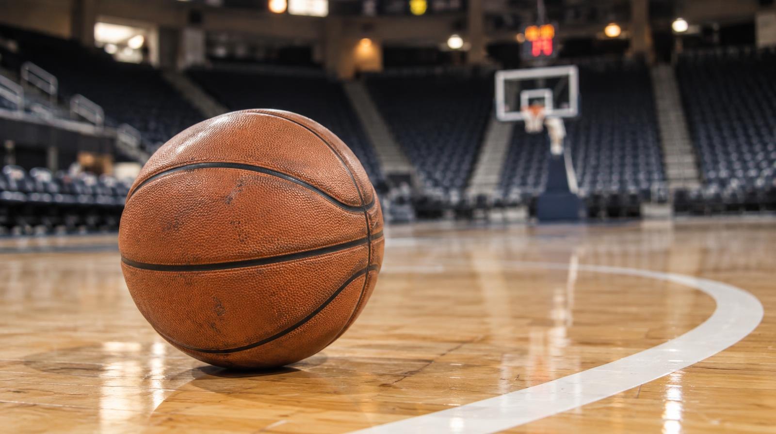 A basketball resting on a hardwood court near the three-point line with empty stadium seating behind it.