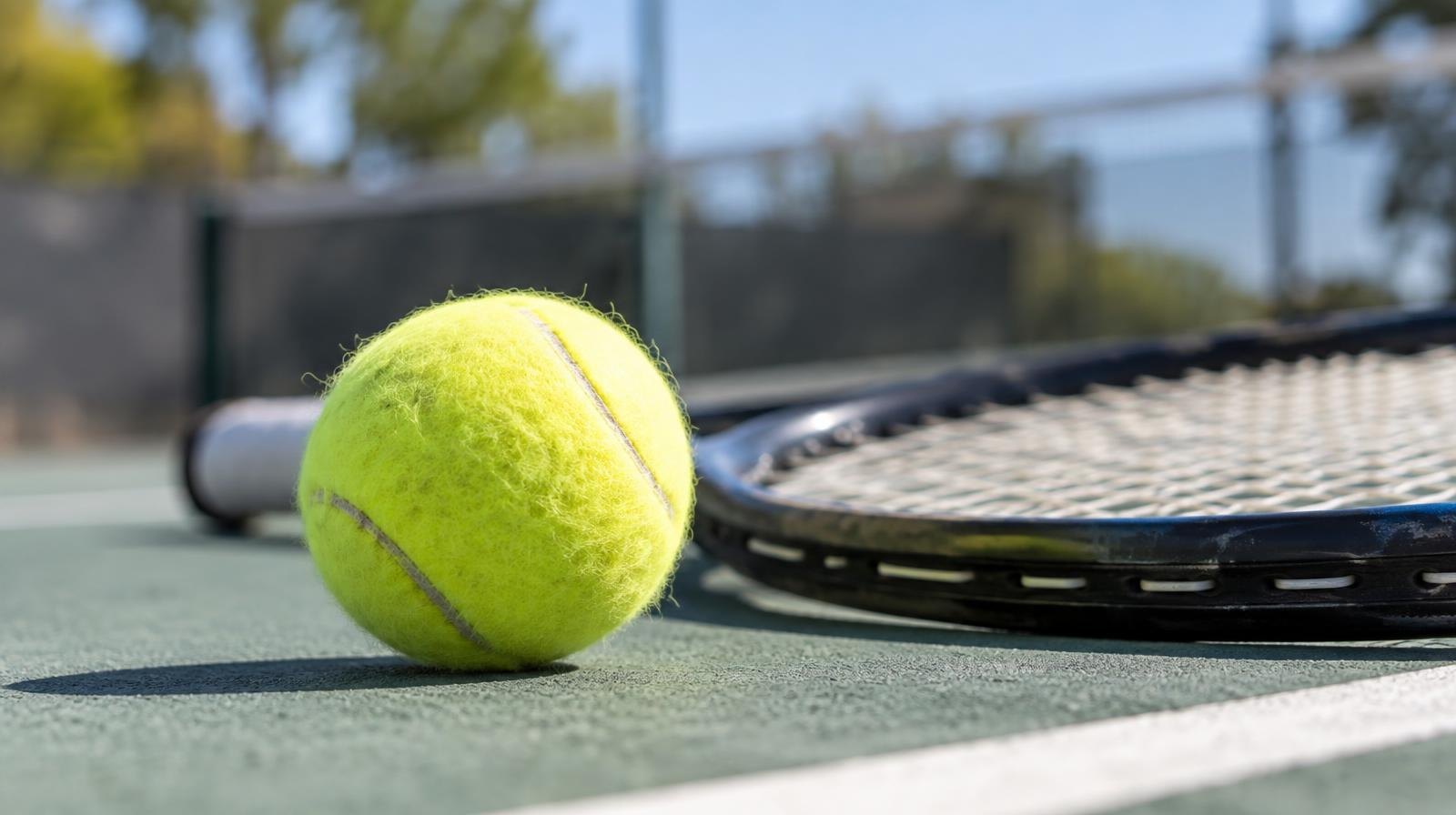 Yellow tennis ball resting beside a tennis racket on a hard court surface.