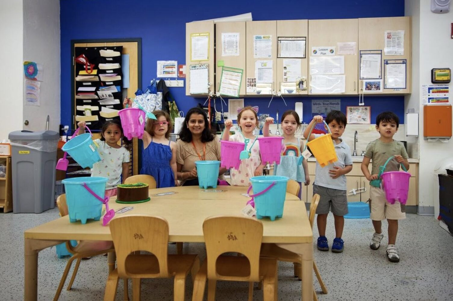 Young children holding colorful buckets stand with two adults in a preschool classroom.
