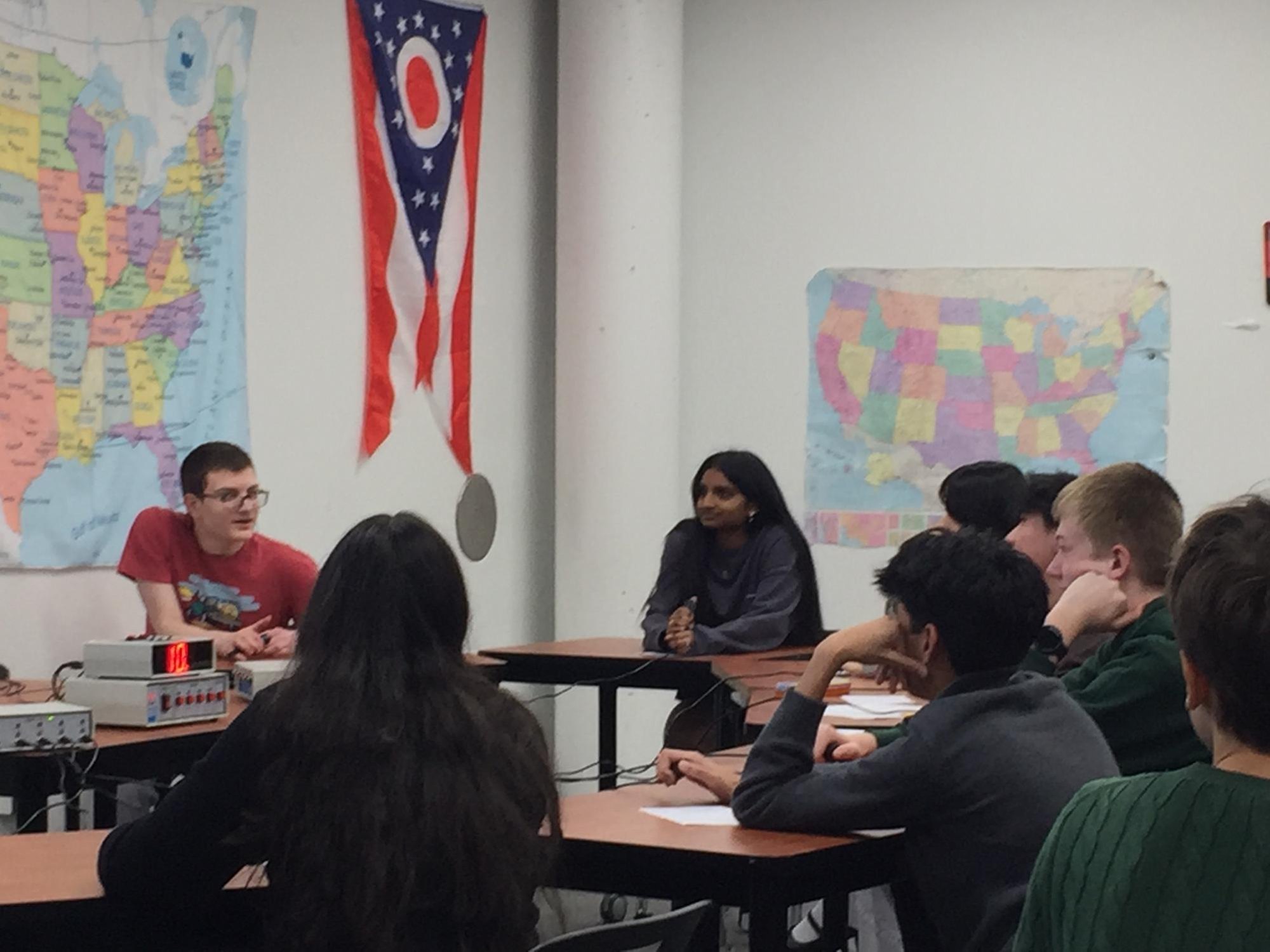 Students seated at classroom desks with a buzzer device visible on a desk nearby.