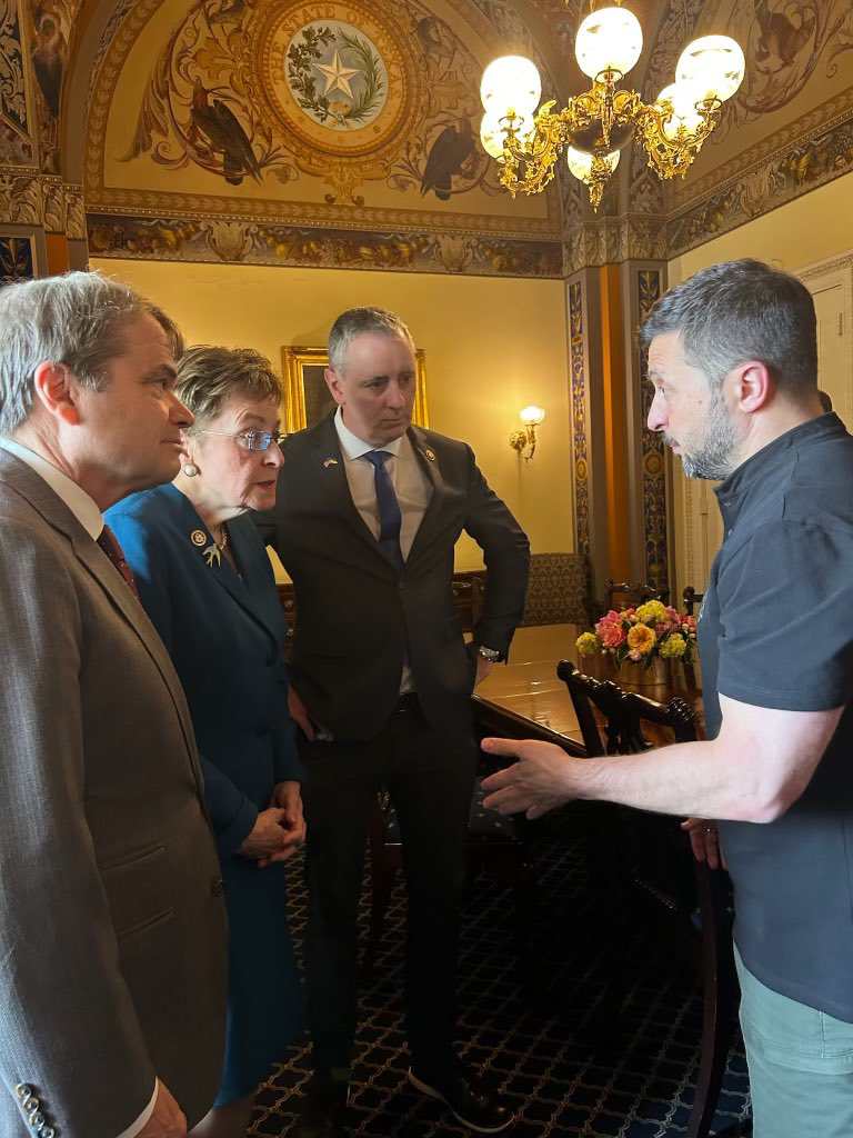 Four people in formal attire conversing in an ornate gilded government room.