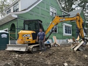 Man in gray t-shirt standing in front of a yellow JCB mini excavator at a residential construction site.