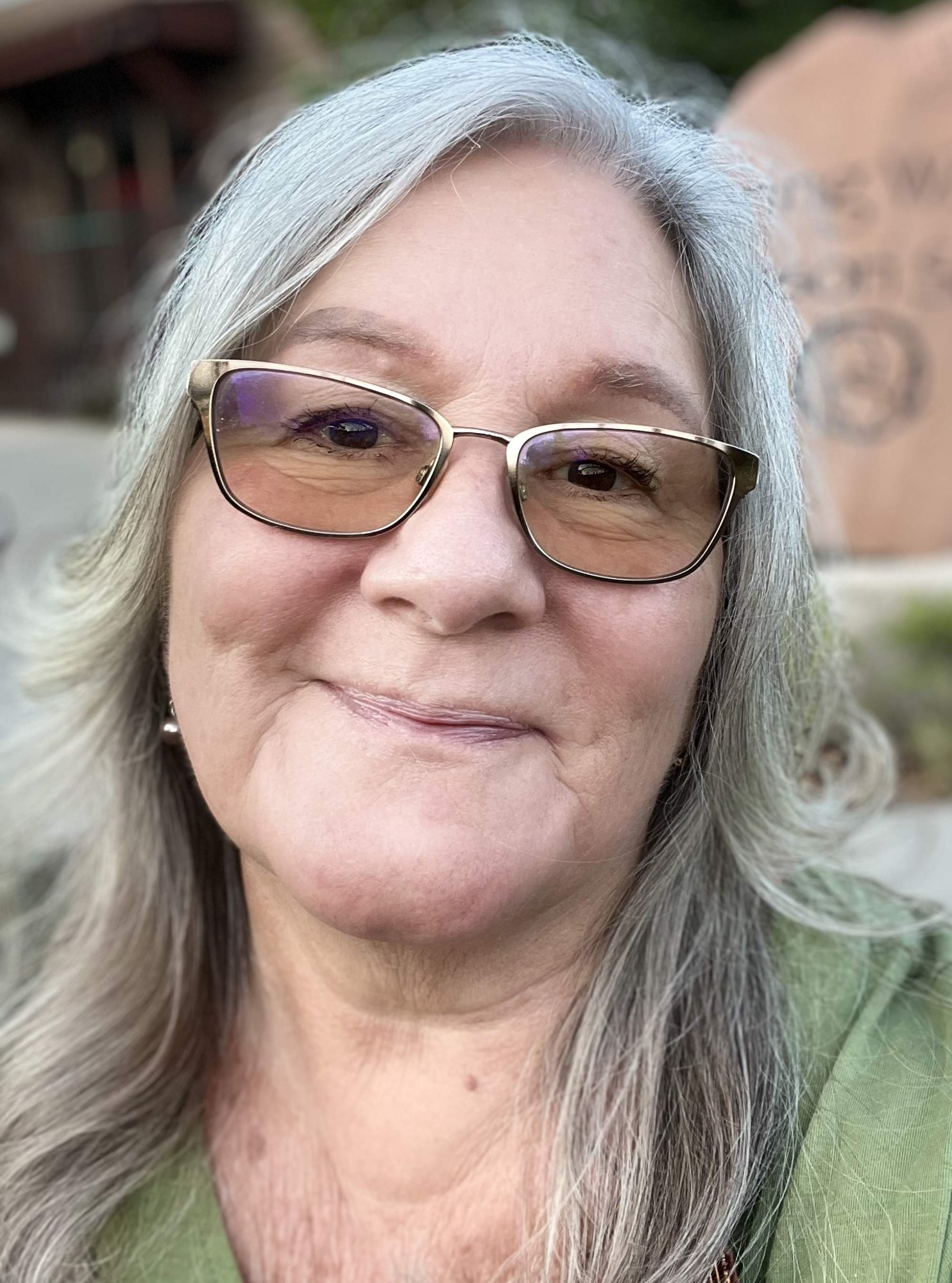 A smiling woman with long gray hair and gold-framed glasses, outdoors near a stone structure.