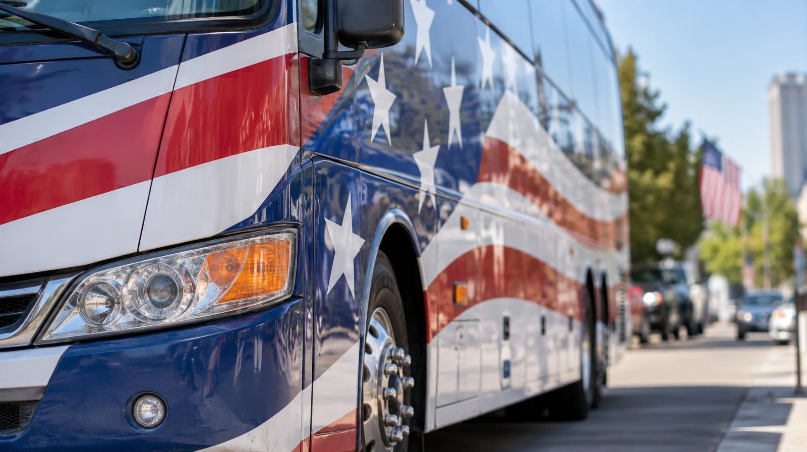 A large campaign bus with red, white, and blue graphics parked on a sunny street.