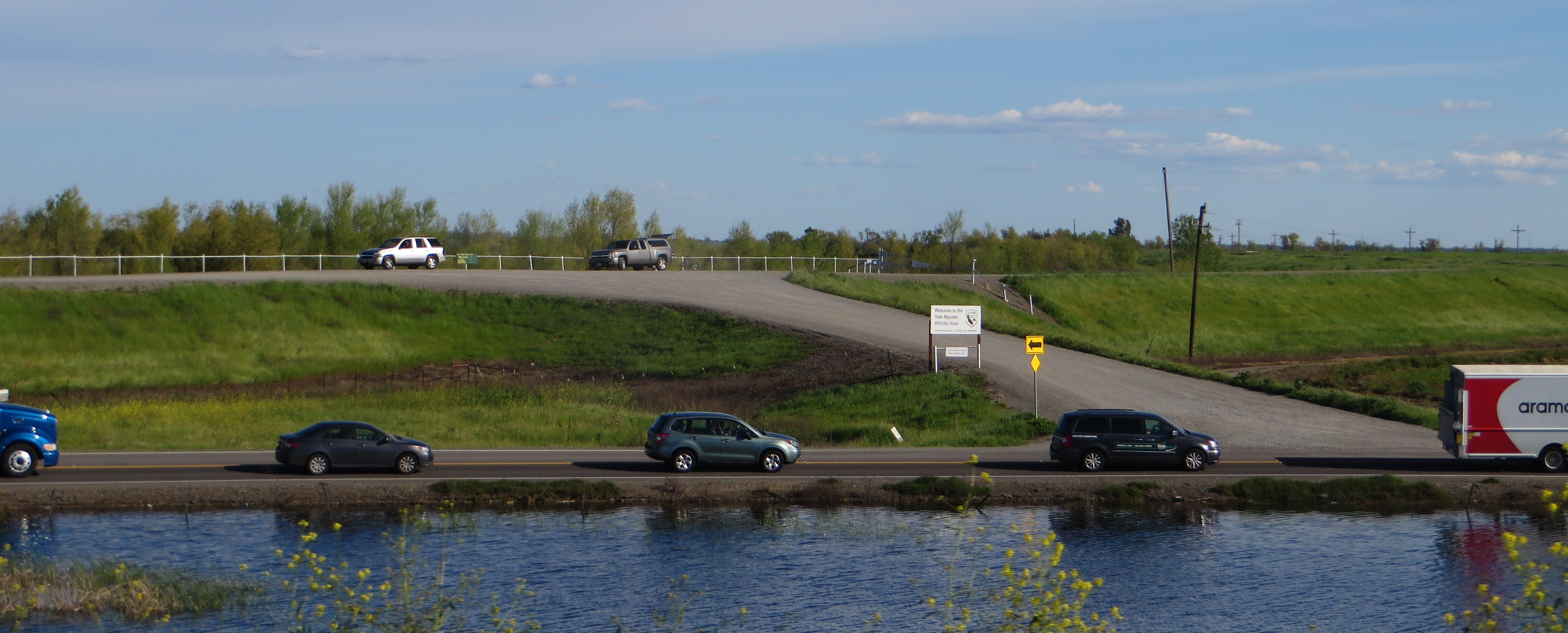 Cars and a truck drive along a road beside standing water near a levee.