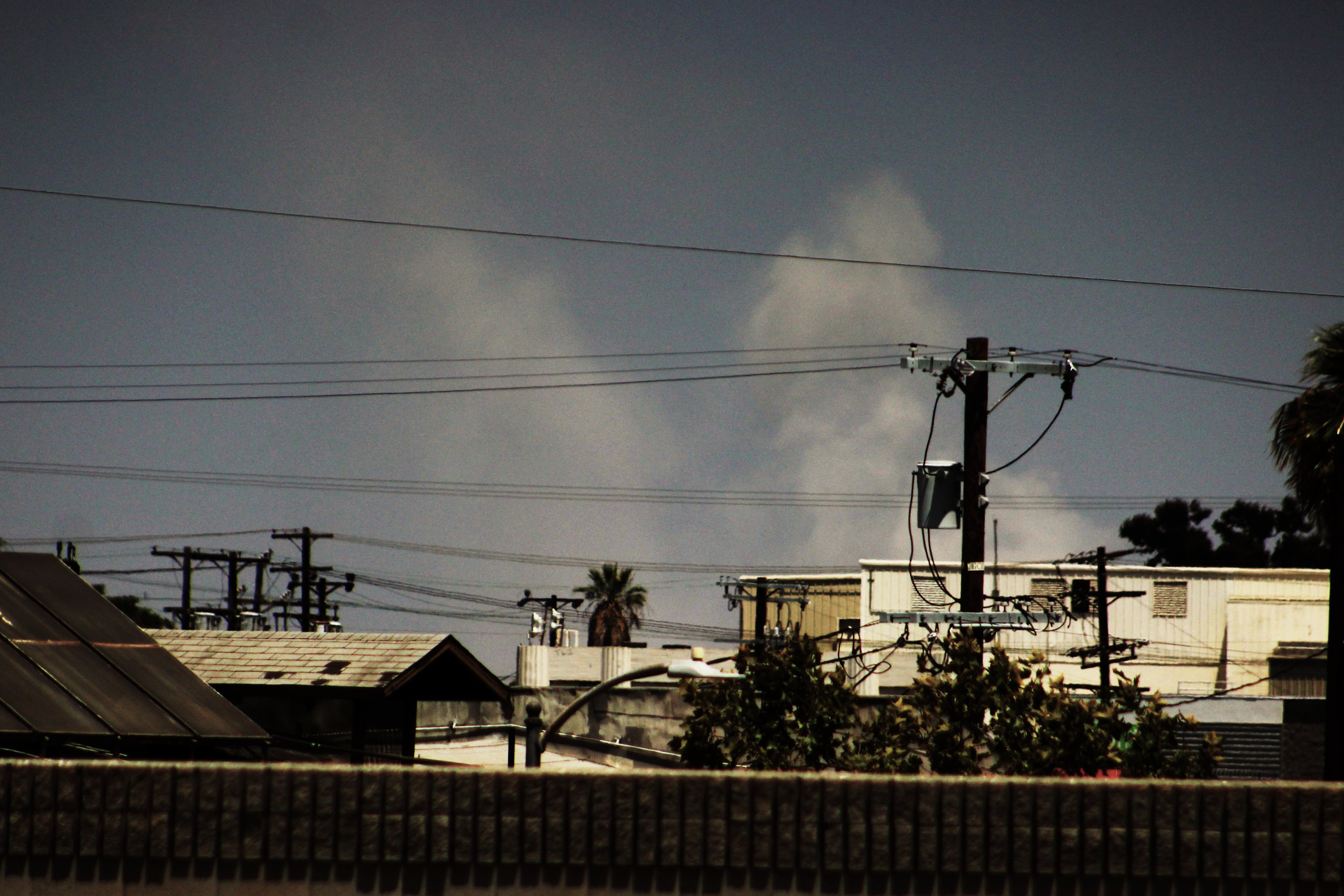 Dark smoke plume rising above rooftops, utility poles, and a palm tree.