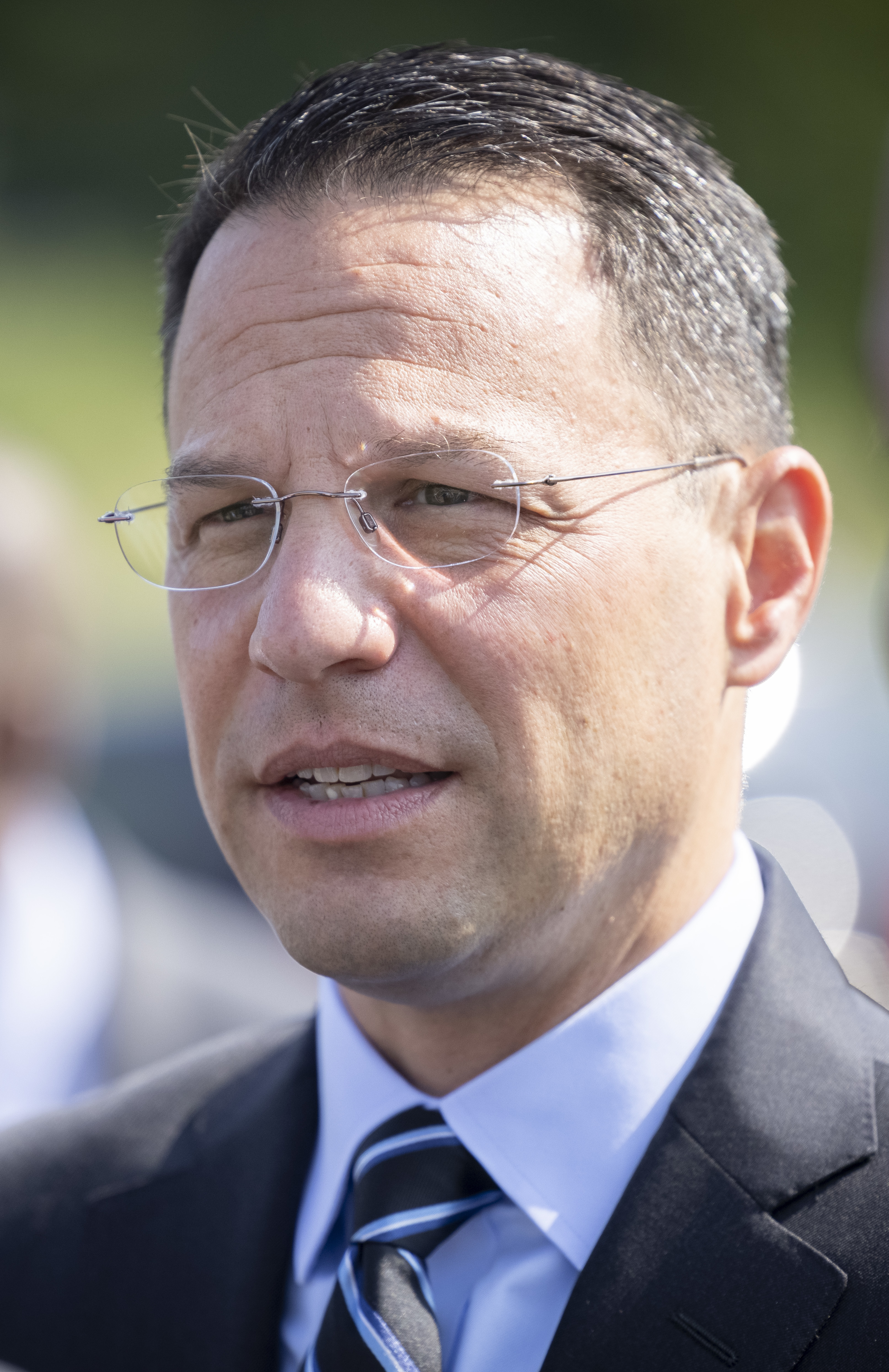 Man in dark suit and wire-rimmed glasses at an outdoor press event.