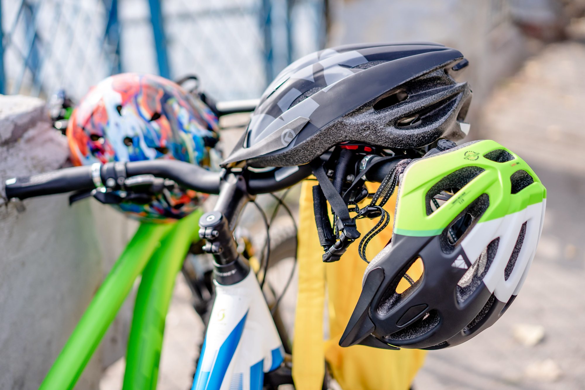 A black adult bike helmet hangs from handlebars next to a green youth helmet and a brightly patterned children's helmet, all clipped to bicycles parked along a fence.