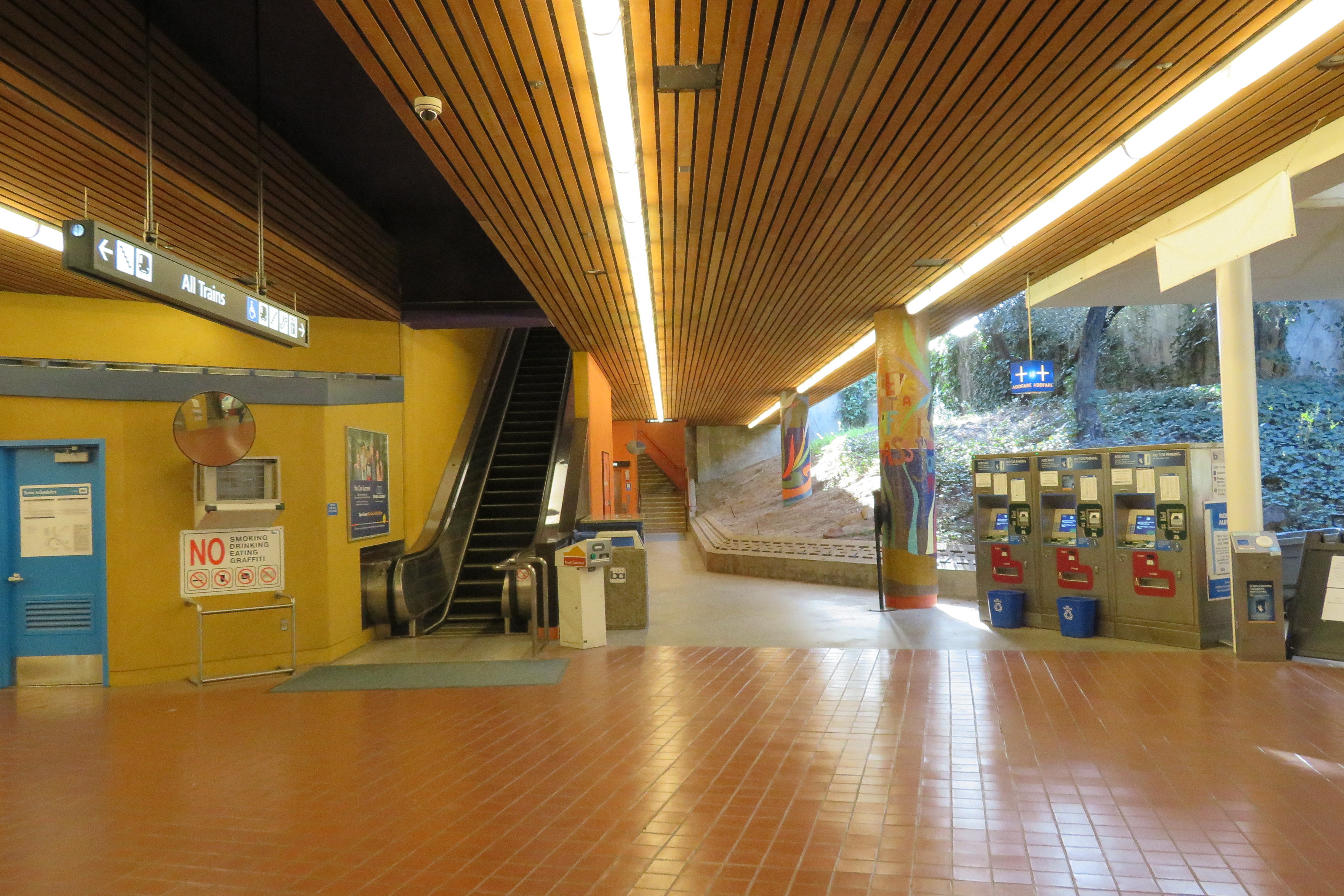 Interior of a transit station fare lobby with ticket vending machines and an escalator.