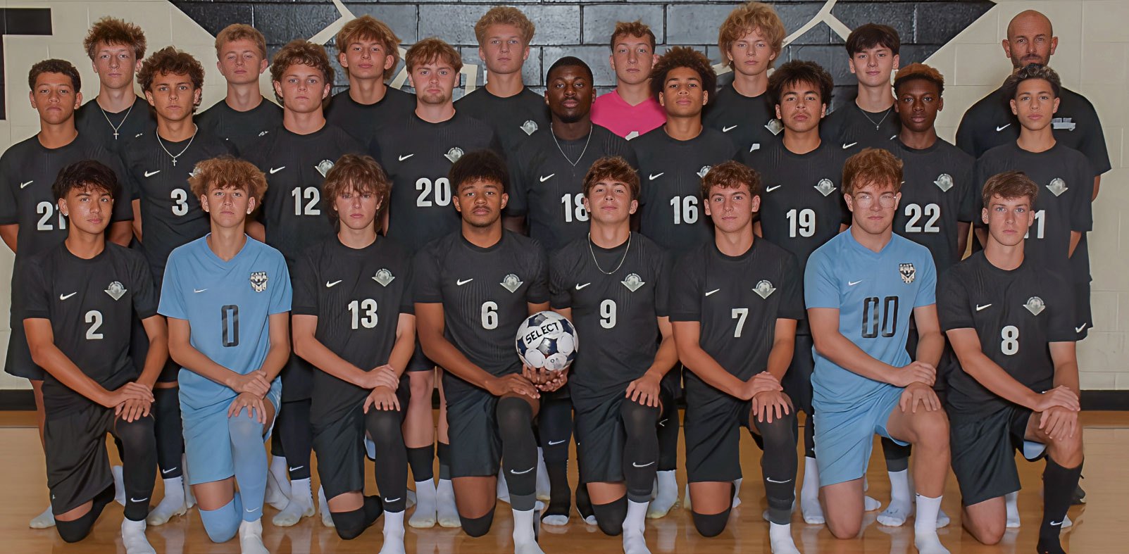 A boys soccer team in dark numbered jerseys poses in two rows for a team photo indoors.