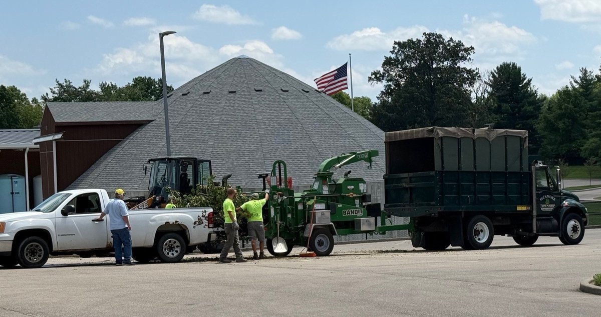 Workers in high-visibility vests operate a green wood chipper between two trucks in a parking lot.