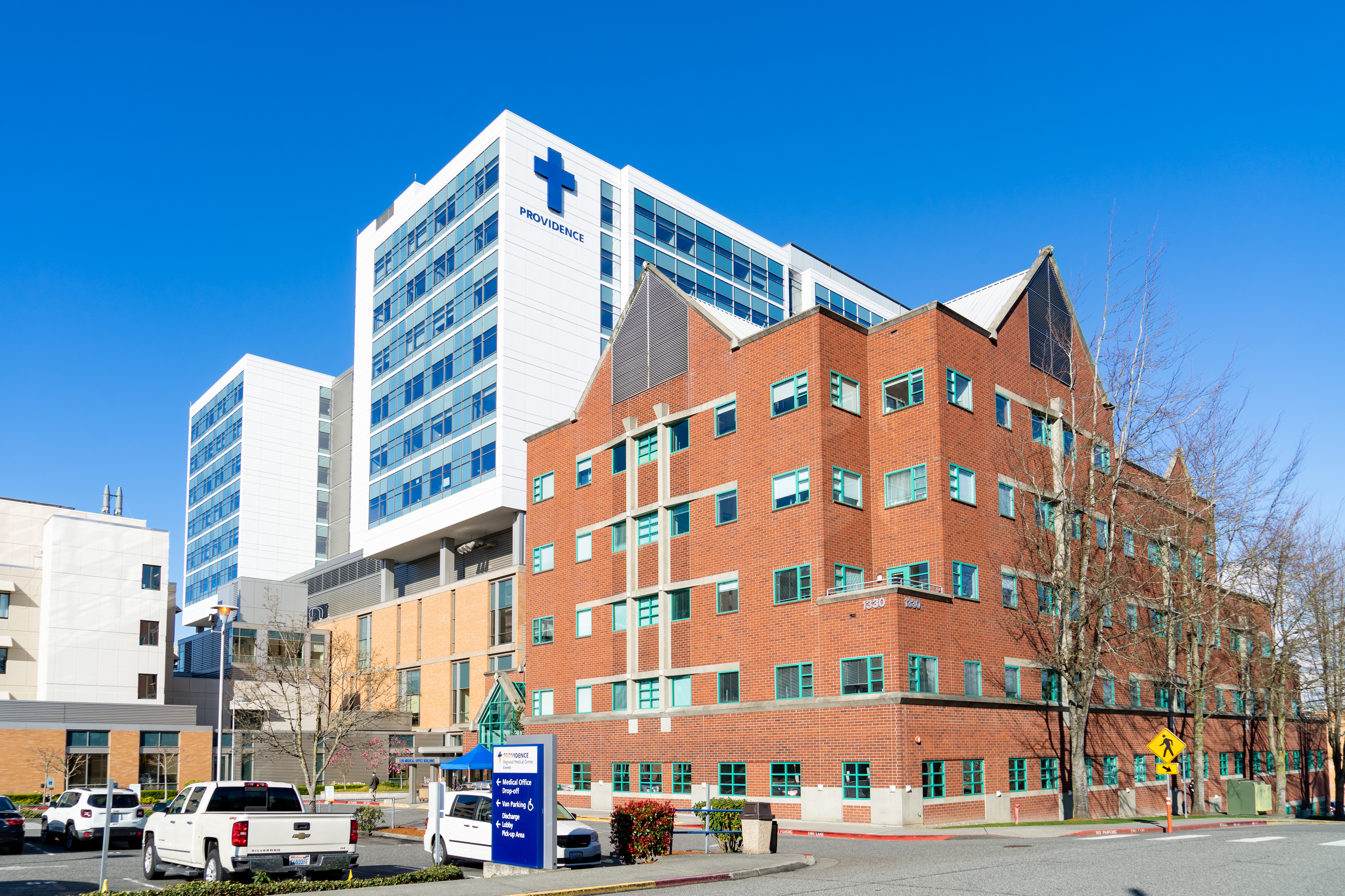 Red-brick hospital building in foreground with a tall white Providence medical tower behind it.