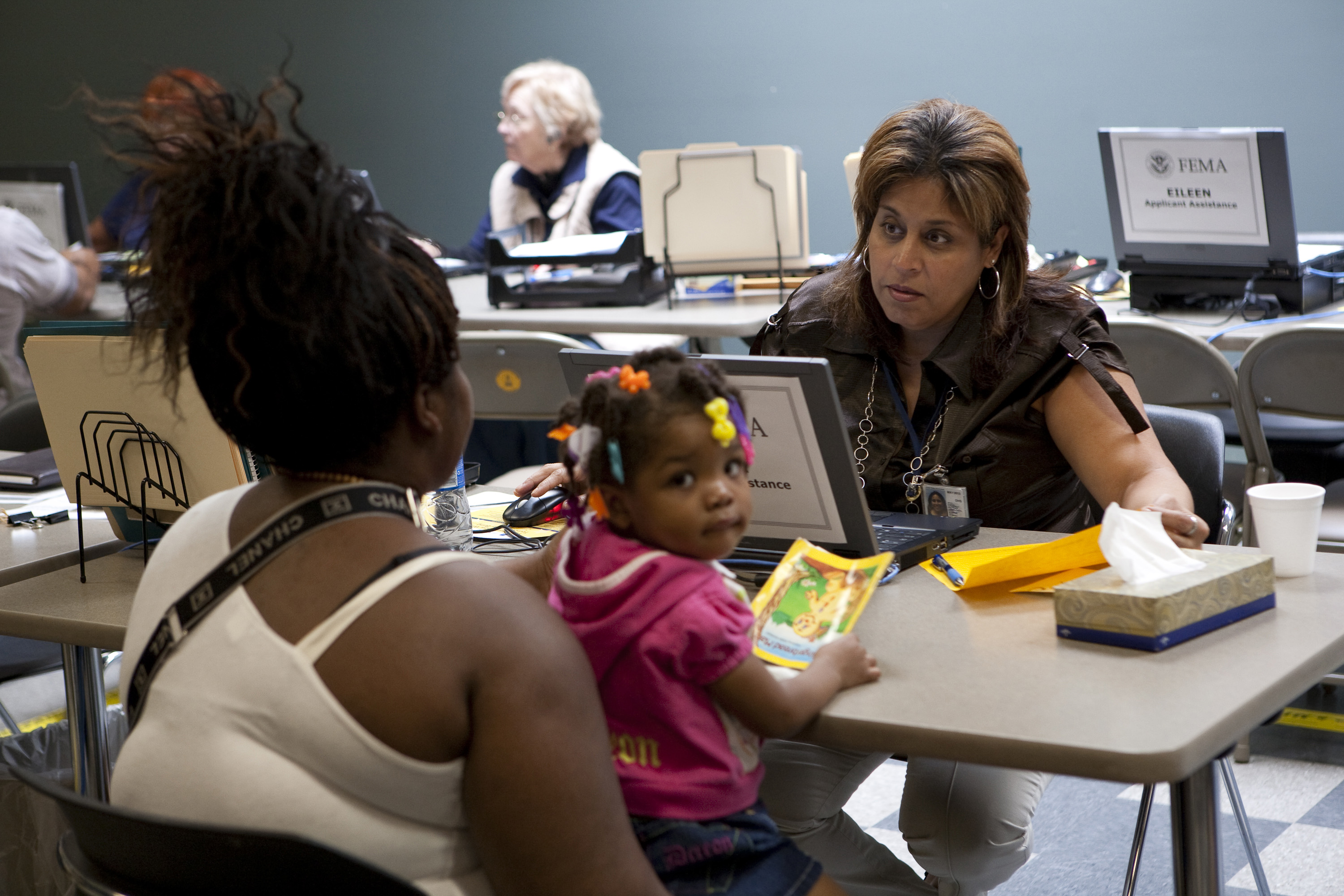 FEMA specialist seated at a desk assists a woman and young child indoors.