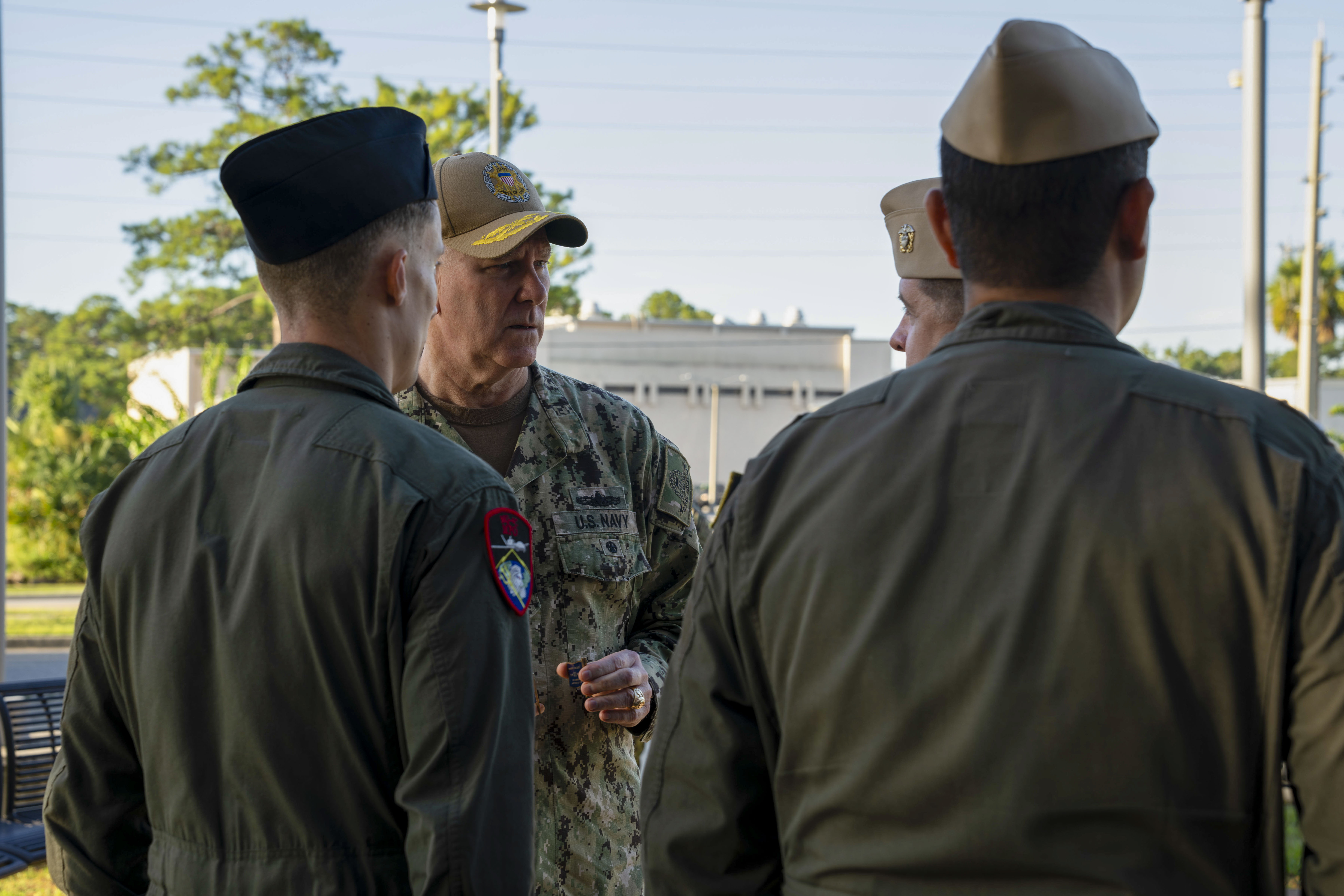 Three uniformed military personnel conversing outdoors at a naval installation with palm trees visible.