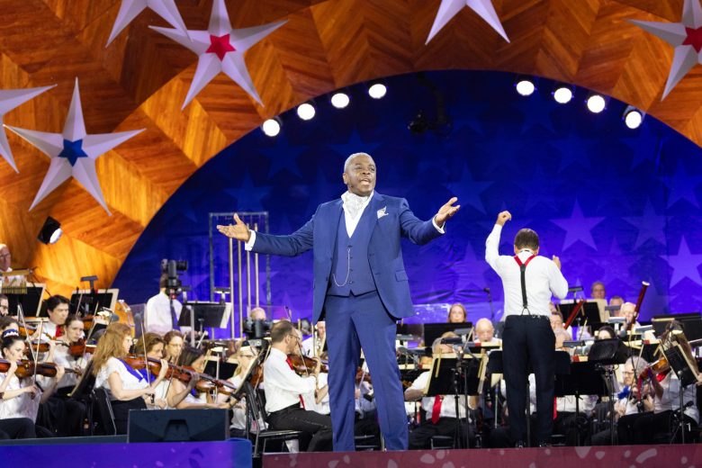 A performer in a blue suit stands center stage at the Hatch Shell, arms outstretched, with a full orchestra behind him.