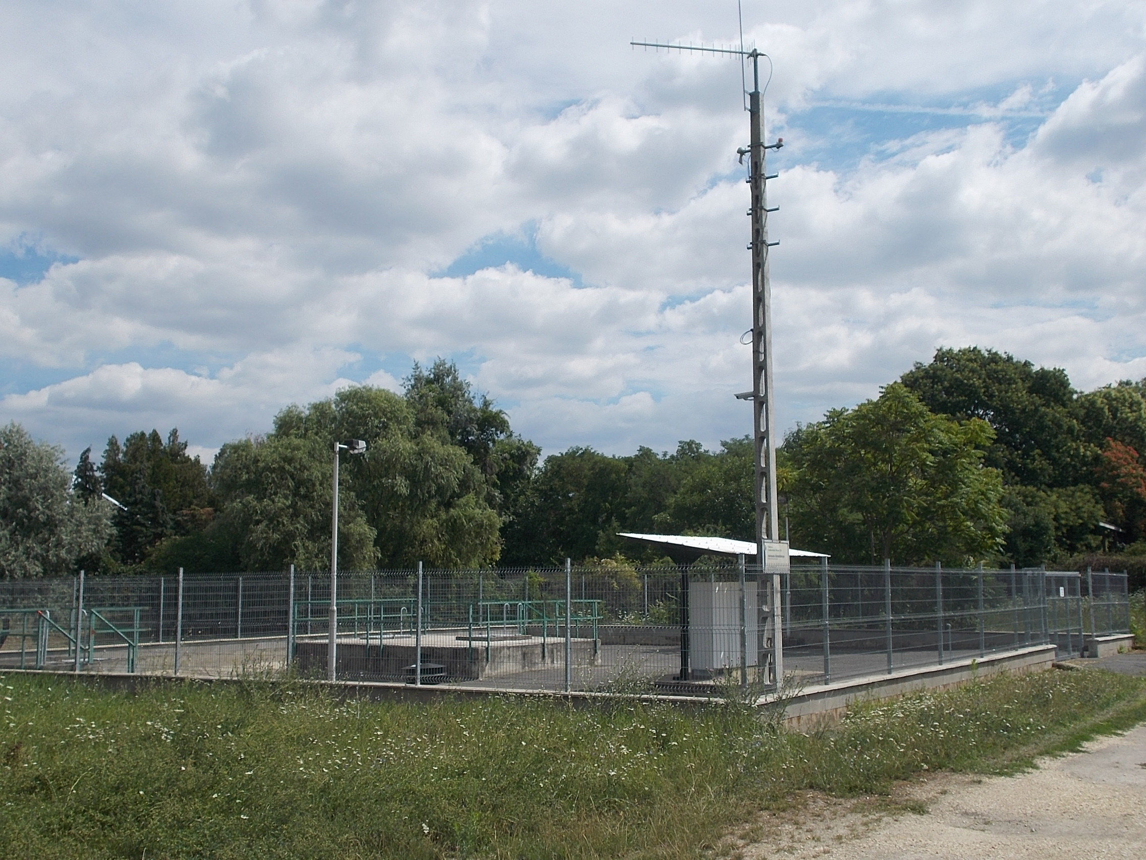 Fenced outdoor water pumping station with green pipes and a metal mast.