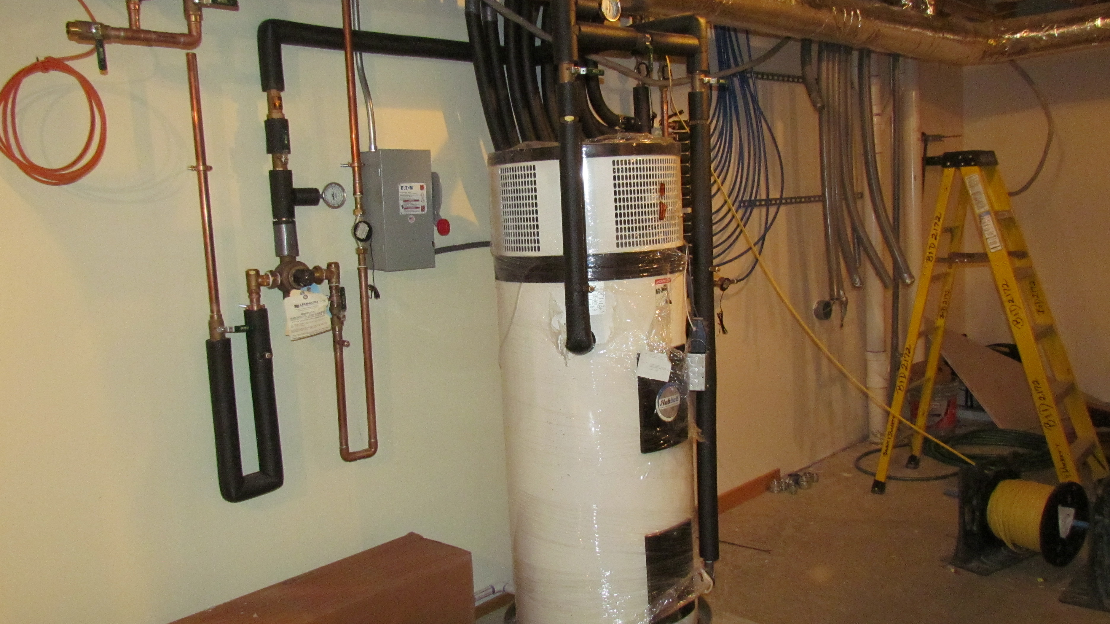 White heat pump water heater tank in a basement utility room with copper pipes.