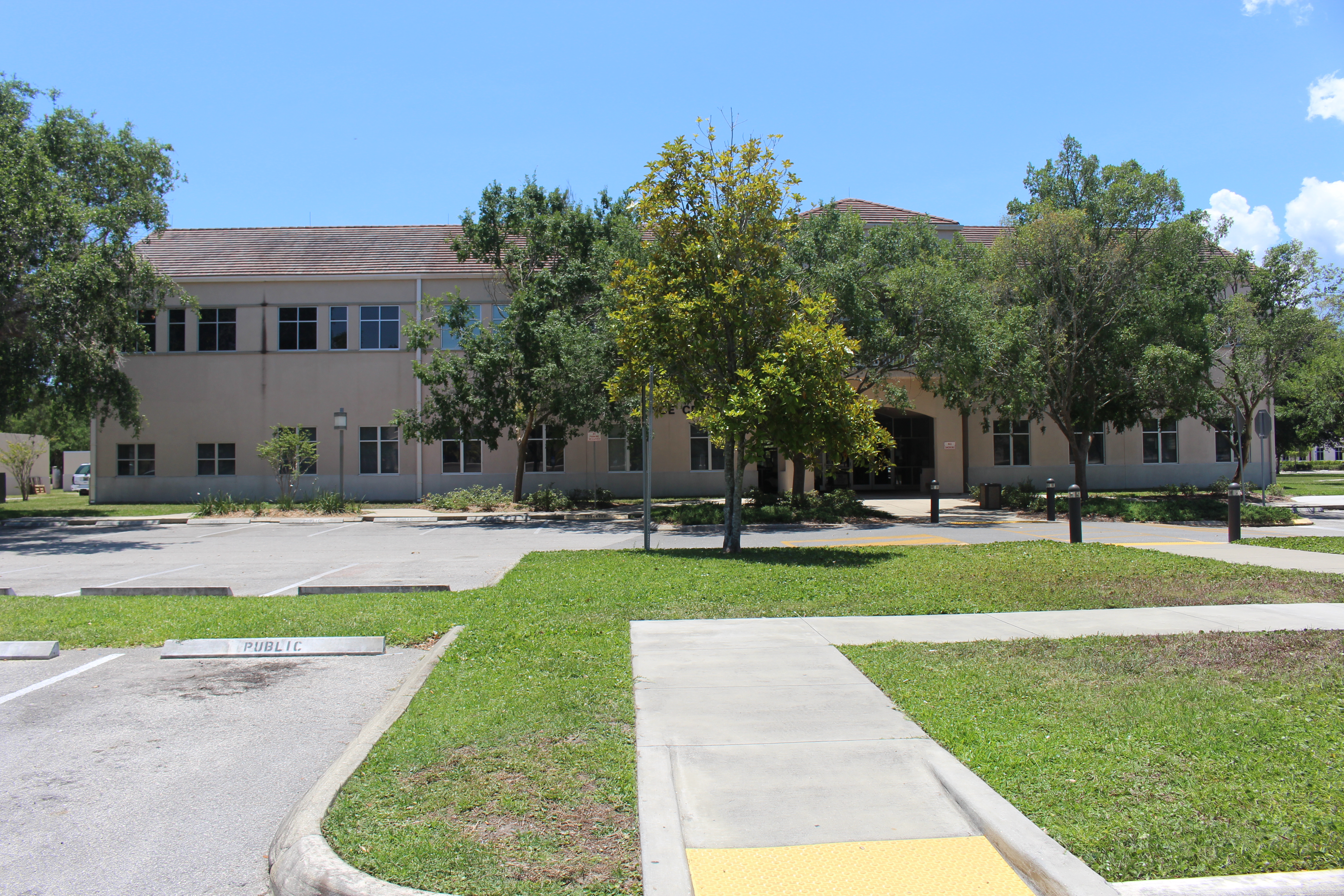 Two-story beige government building with trees and a parking lot in front.