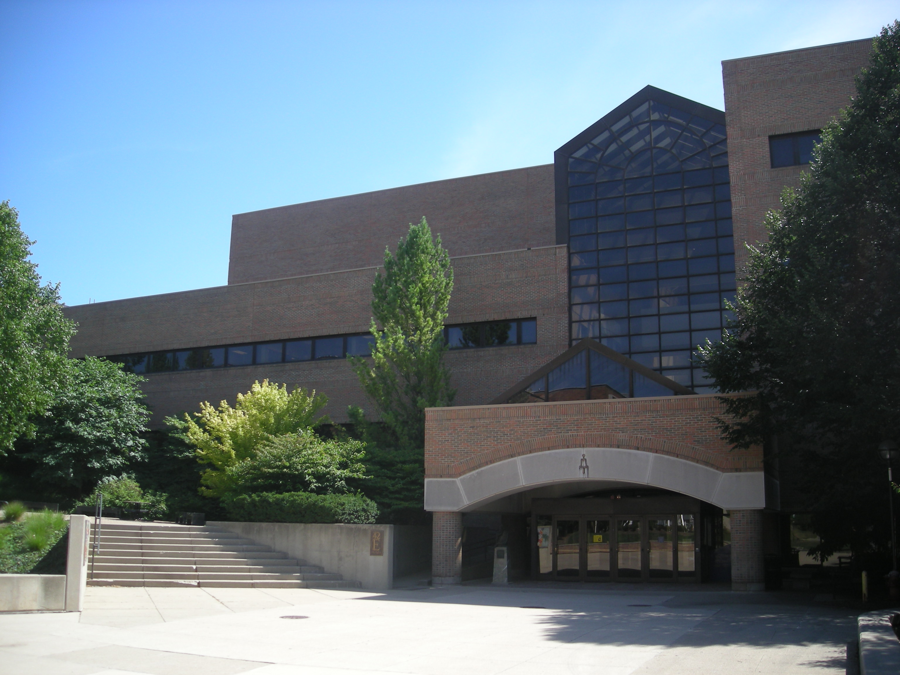 Brick and glass university building with a covered entrance on a tree-lined campus plaza.