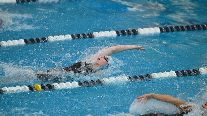 A swimmer performs backstroke in an indoor competition pool with lane dividers visible.