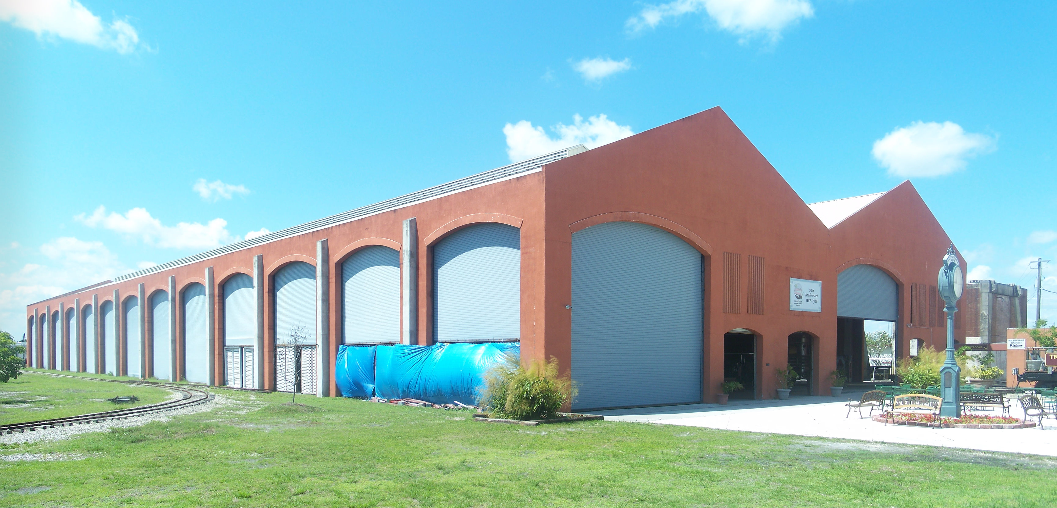 Exterior of a large red brick building with arched openings and railroad tracks in the foreground.