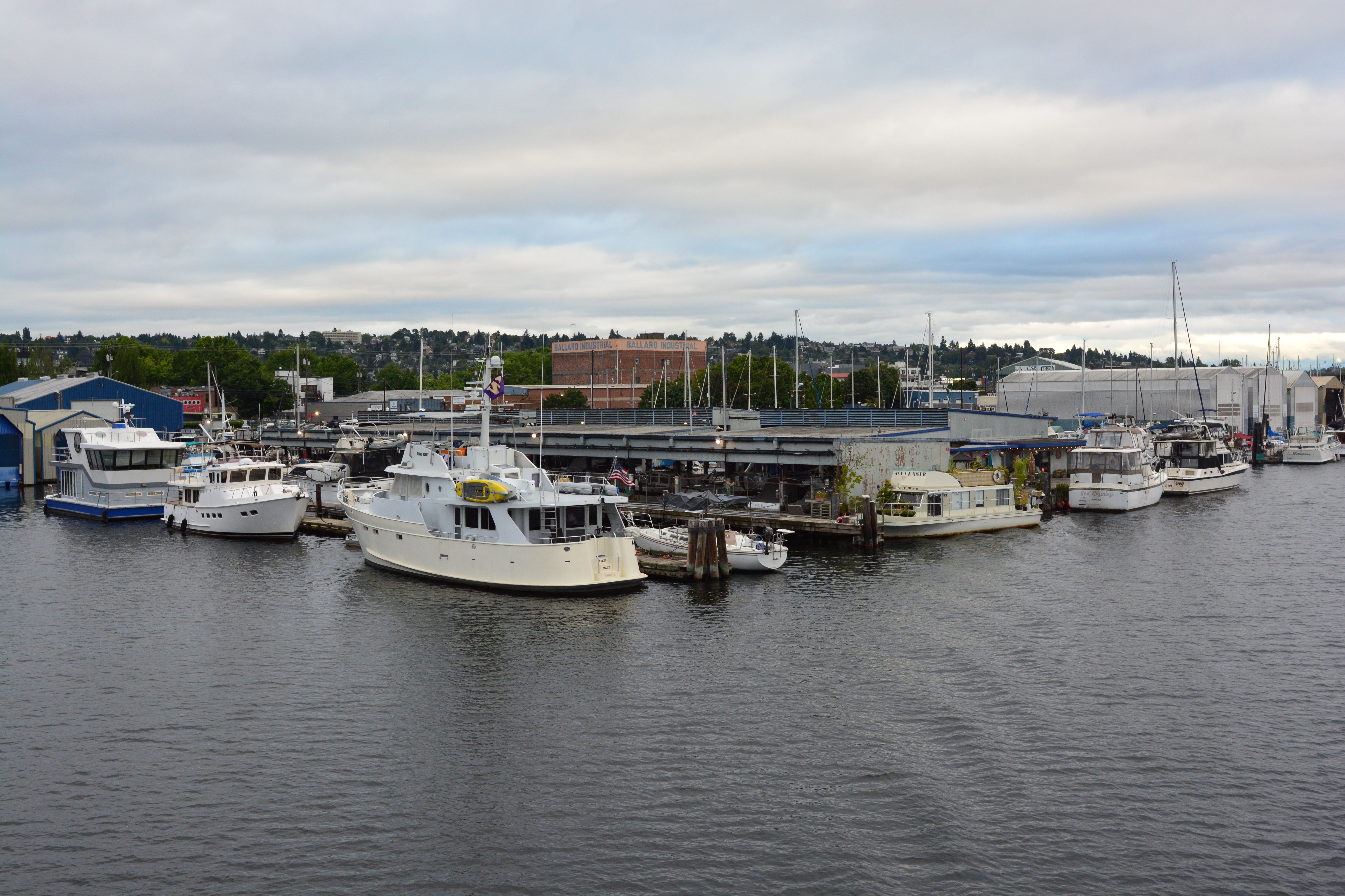 Boats docked at a marina on calm water with industrial waterfront buildings behind.