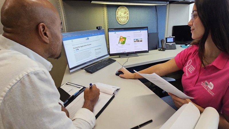 A woman in a pink polo and a man with a clipboard sit at a desk with two computer monitors.